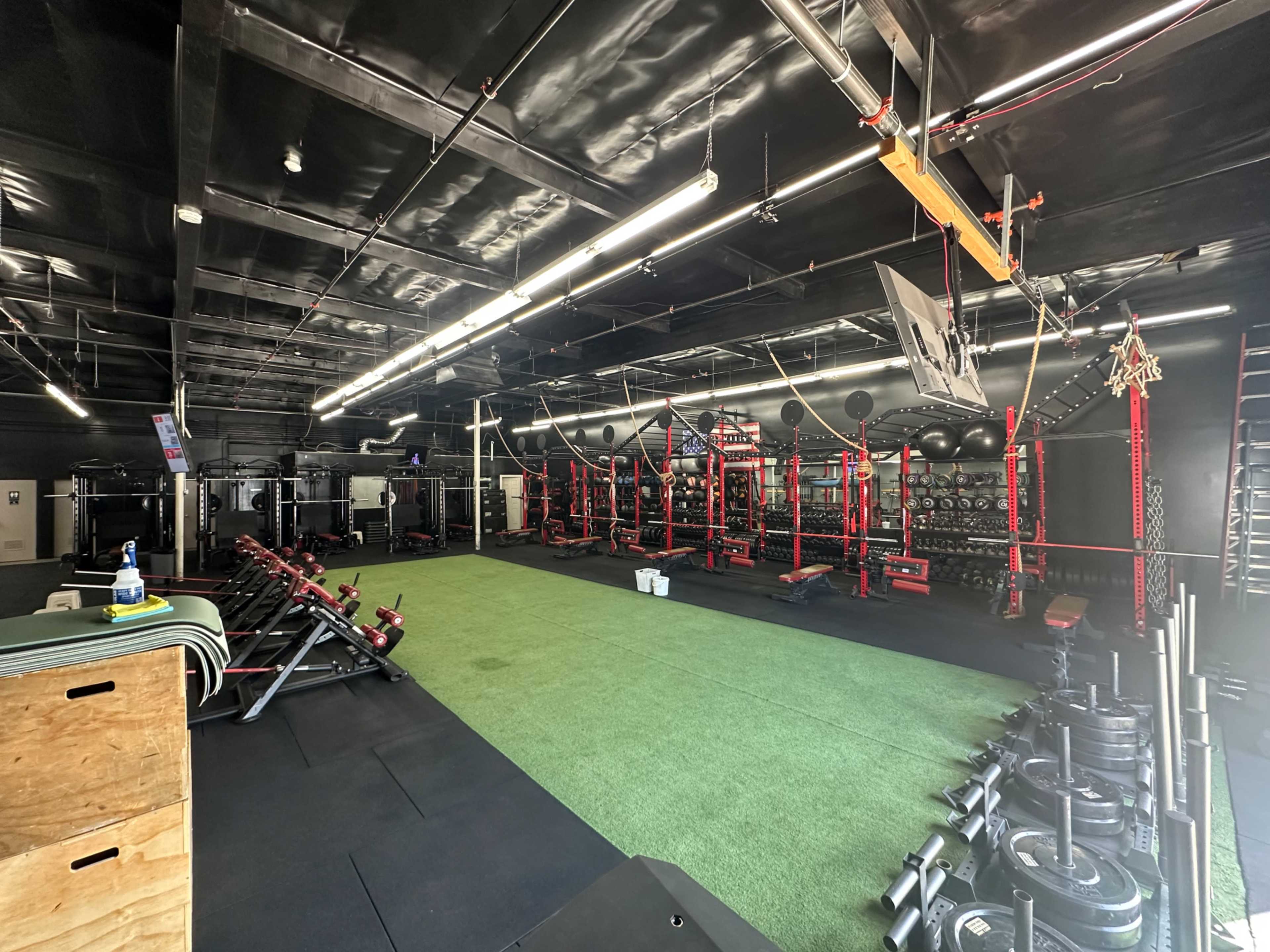 The image shows a well-equipped gym with black and red workout stations, a turf area, and various weights and fitness equipment arranged neatly.