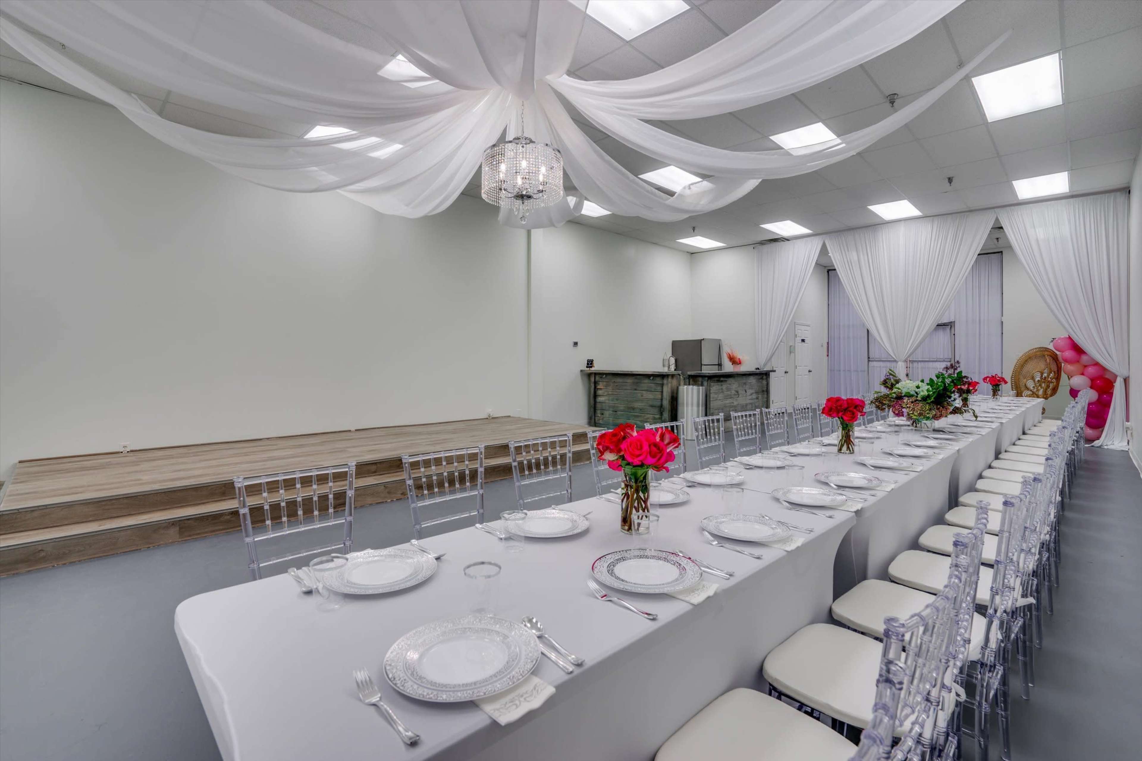 A long banquet table set with white tablecloths, clear chairs, and floral centerpieces is arranged in a spacious room with draped fabric overhead and a small stage at one end.