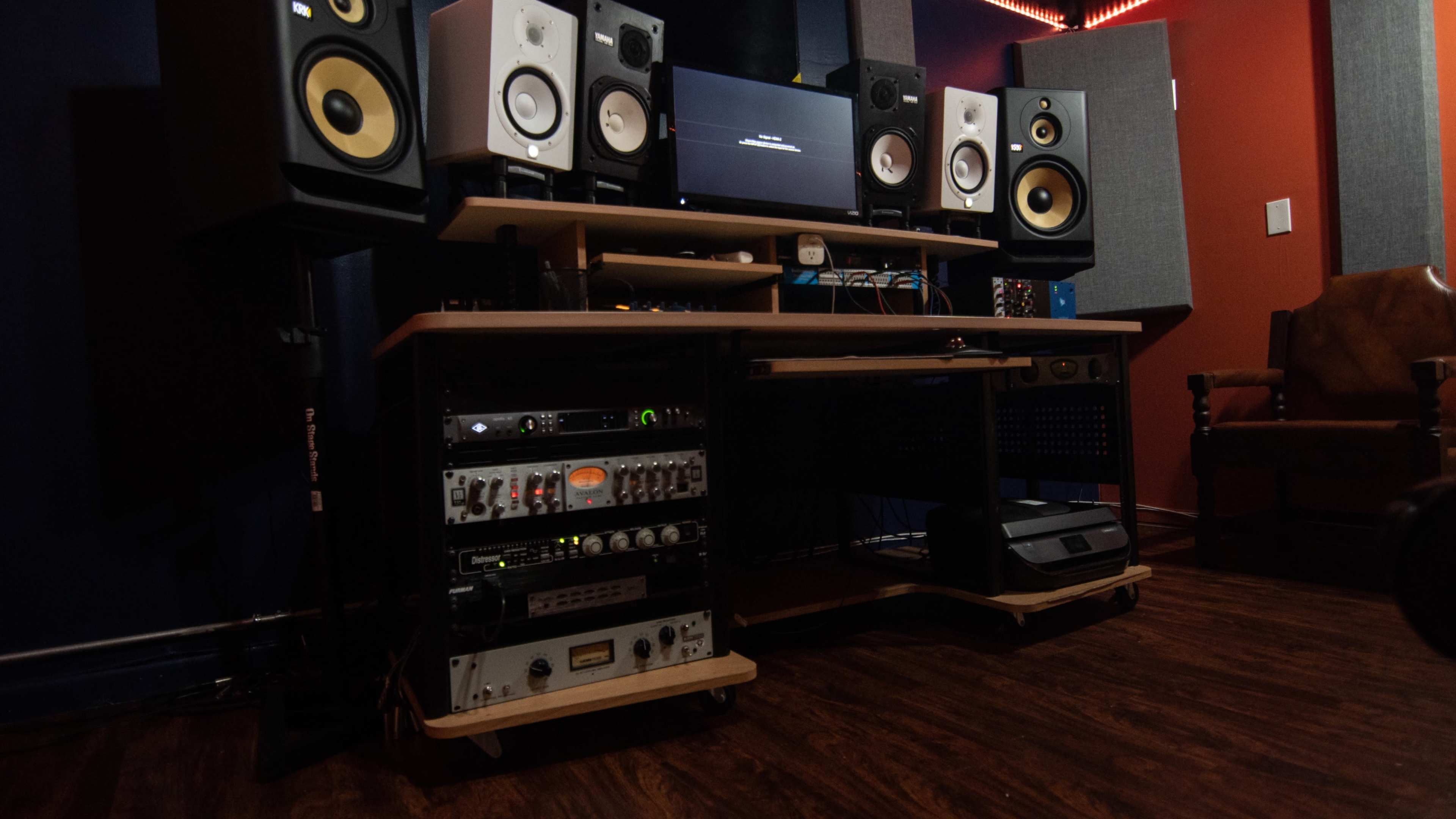 The image shows a music studio setup with speaker monitors, a computer screen, and various audio equipment arranged on a desk.