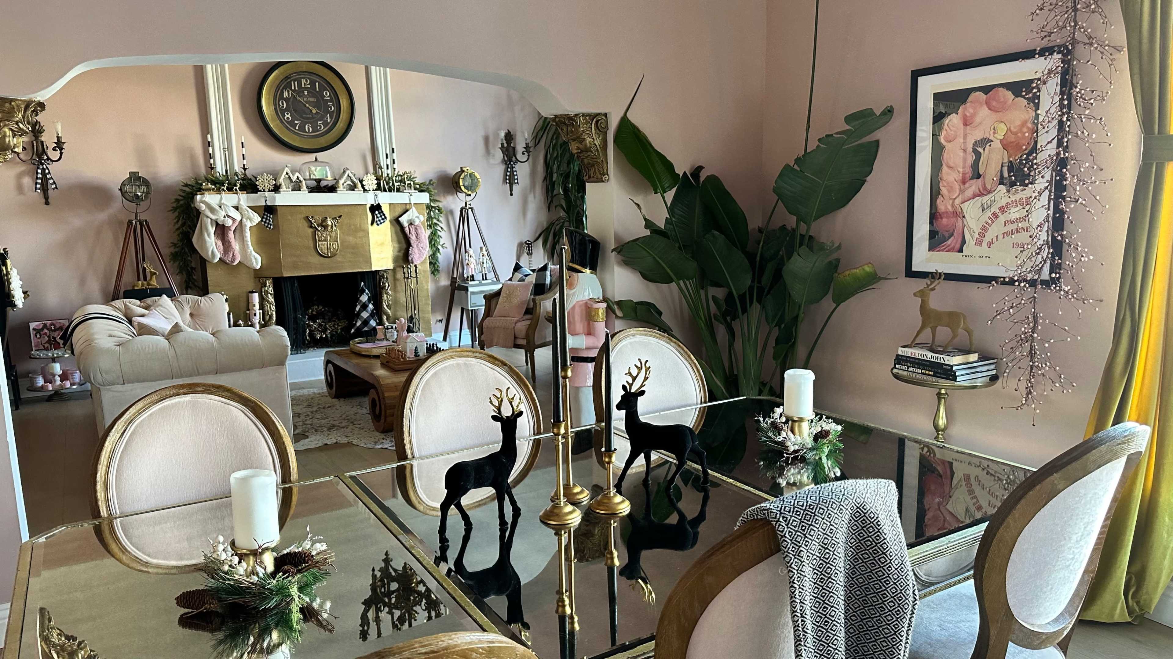 The image shows a styled dining area with a glass table, gold candlesticks, and decorative reindeer, overlooking a living room with a fireplace adorned for the holidays.