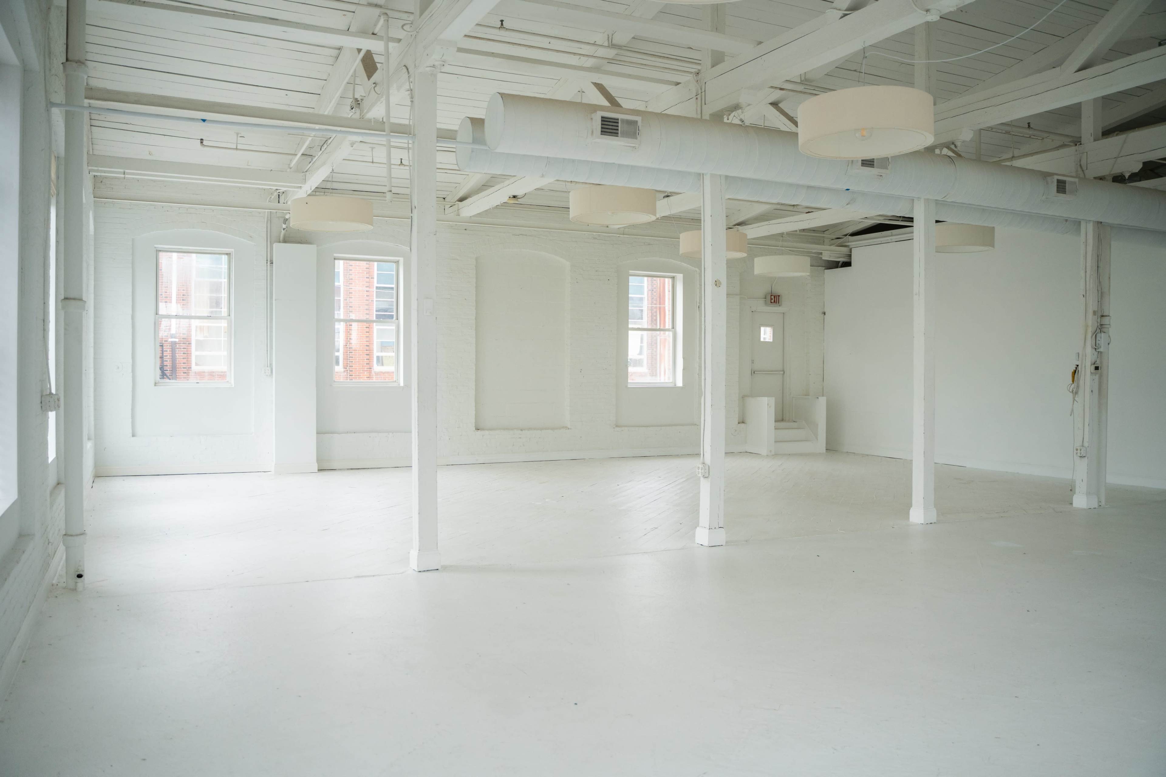 The image shows a spacious, white-walled room with large windows, exposed beams, and a polished floor.