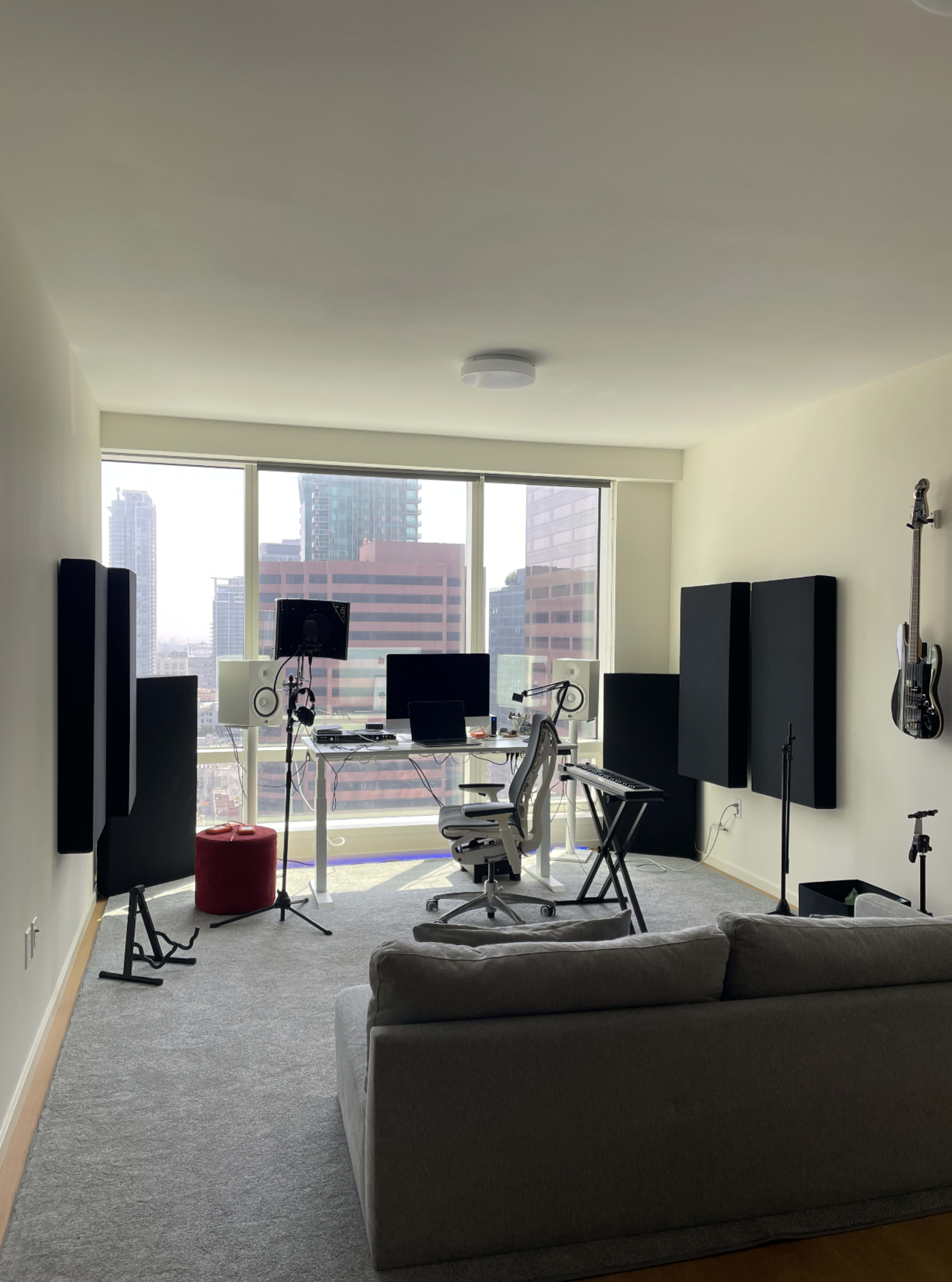 A modern home office setup features a clear desk with a computer, chairs, and soundproof panels, all situated in a well-lit room with large windows overlooking a city skyline.