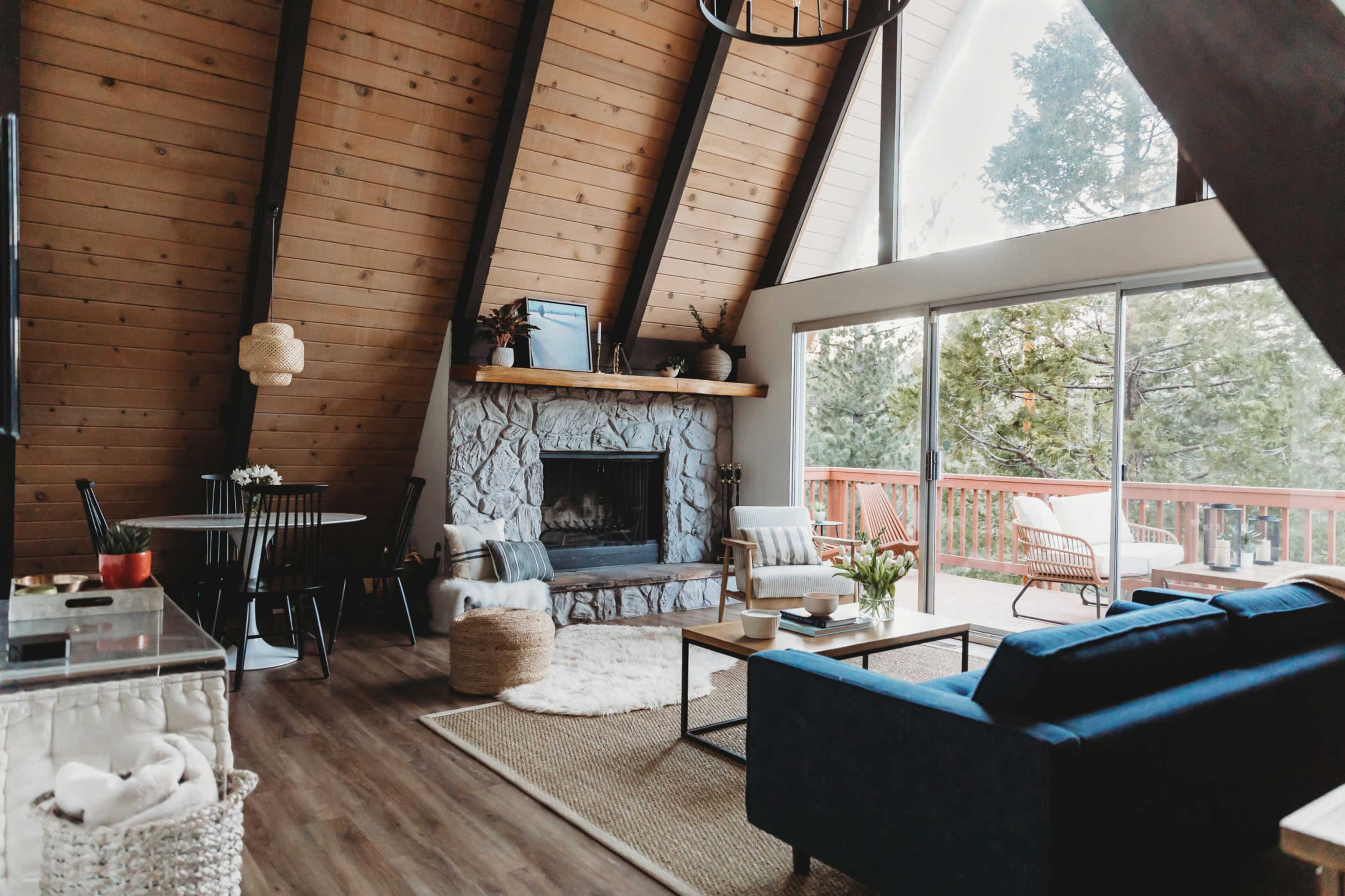 A cozy, A-frame living room with a stone fireplace, large windows, and wooden beams, featuring a blue sofa and a table set for dining.
