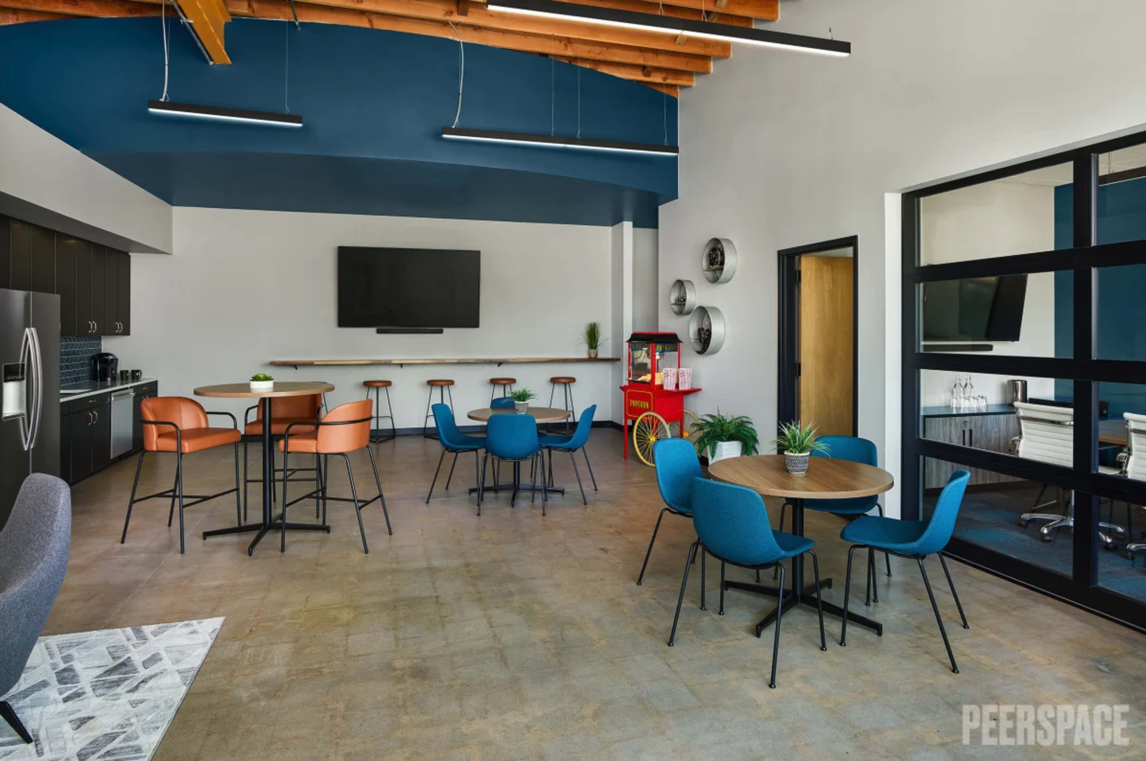 A modern, open-plan office break room featuring a kitchen area, various seating arrangements, and a large wall-mounted TV.