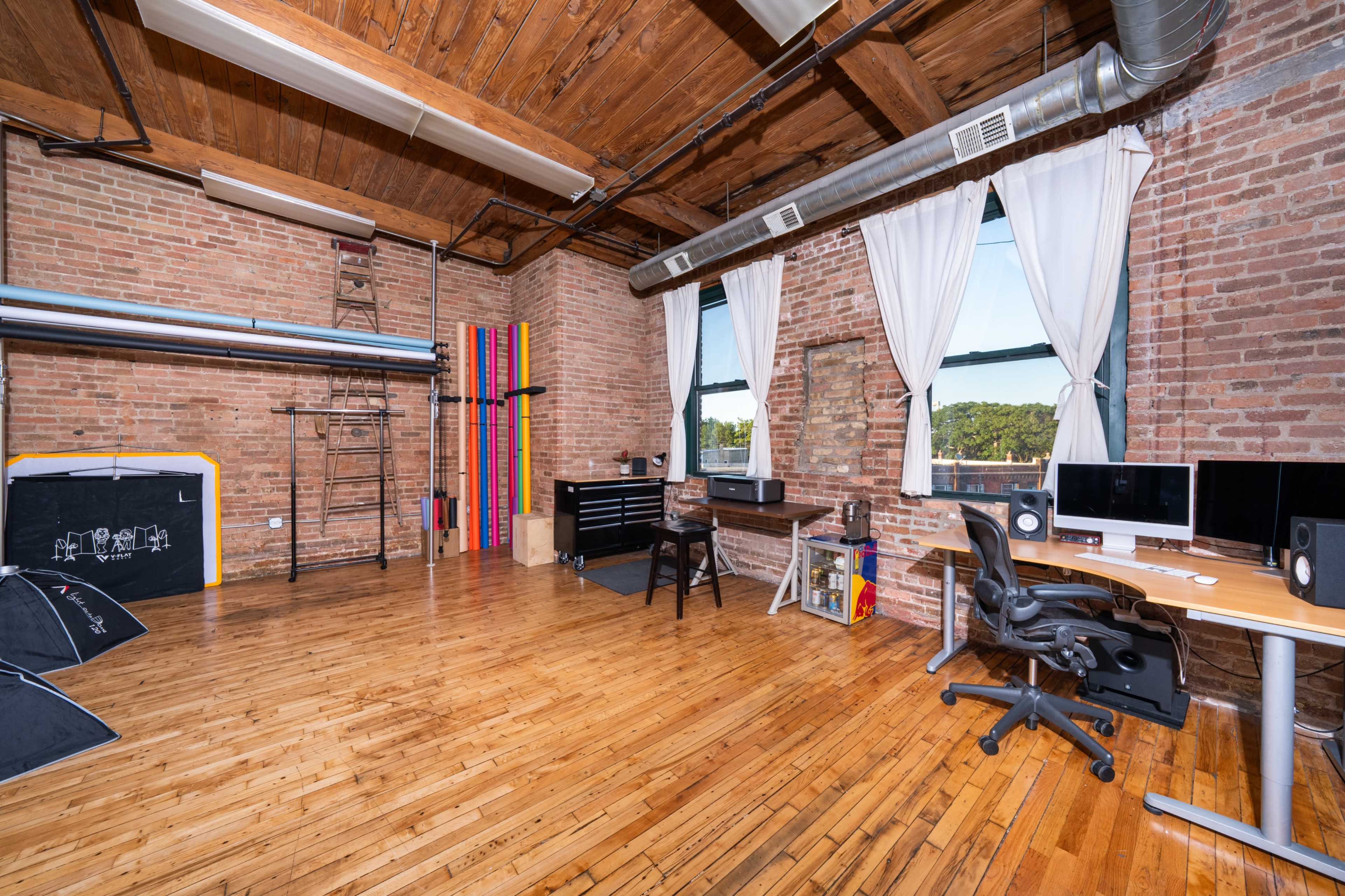 The image shows a spacious office with wooden floors, exposed brick walls, large windows, and various office furniture, including a desk, computer, and a set of colorful gymnastic equipment.