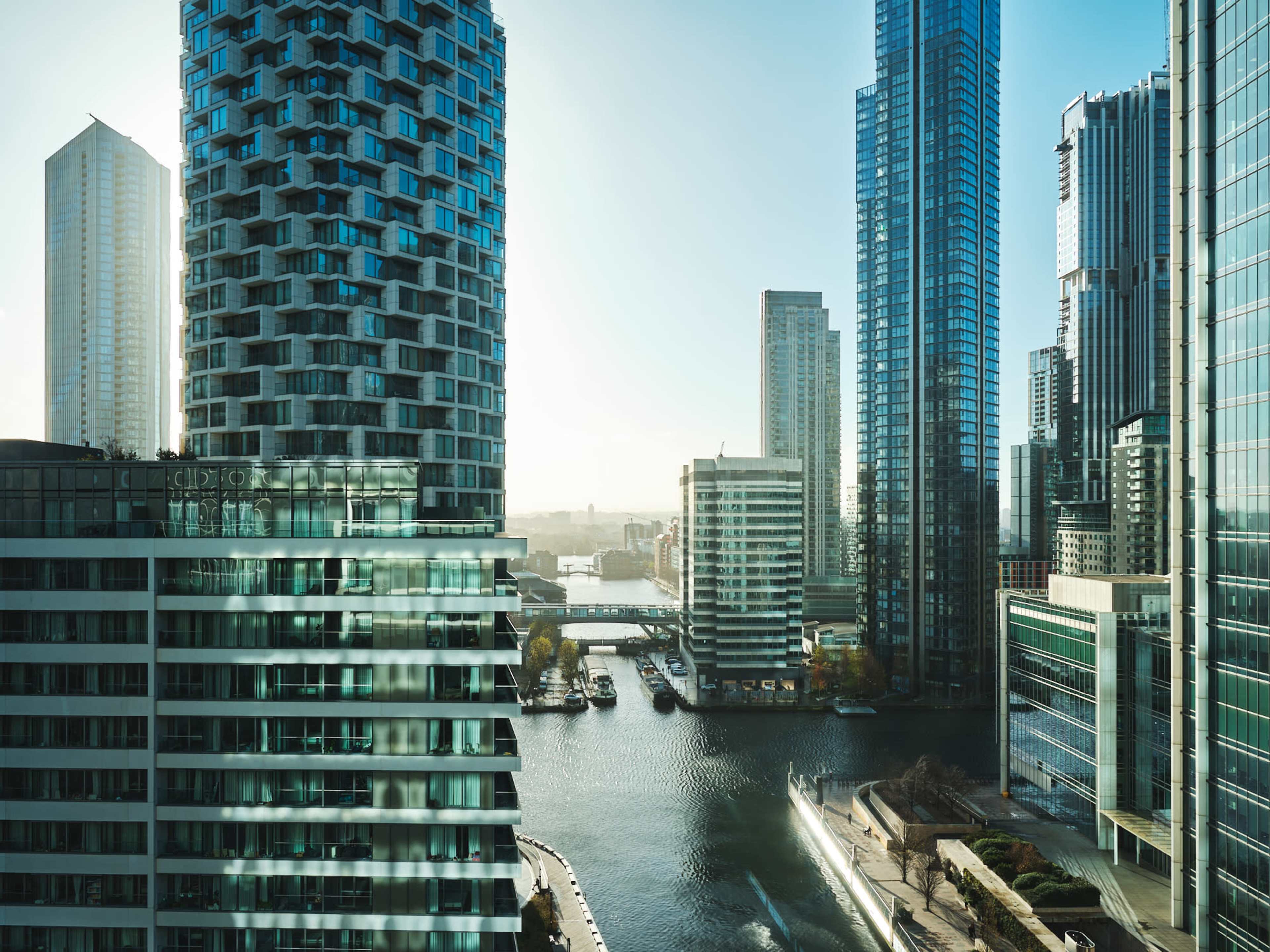 A view of modern skyscrapers along a waterfront in a city, with reflections on the water and boats docked along the shore.
