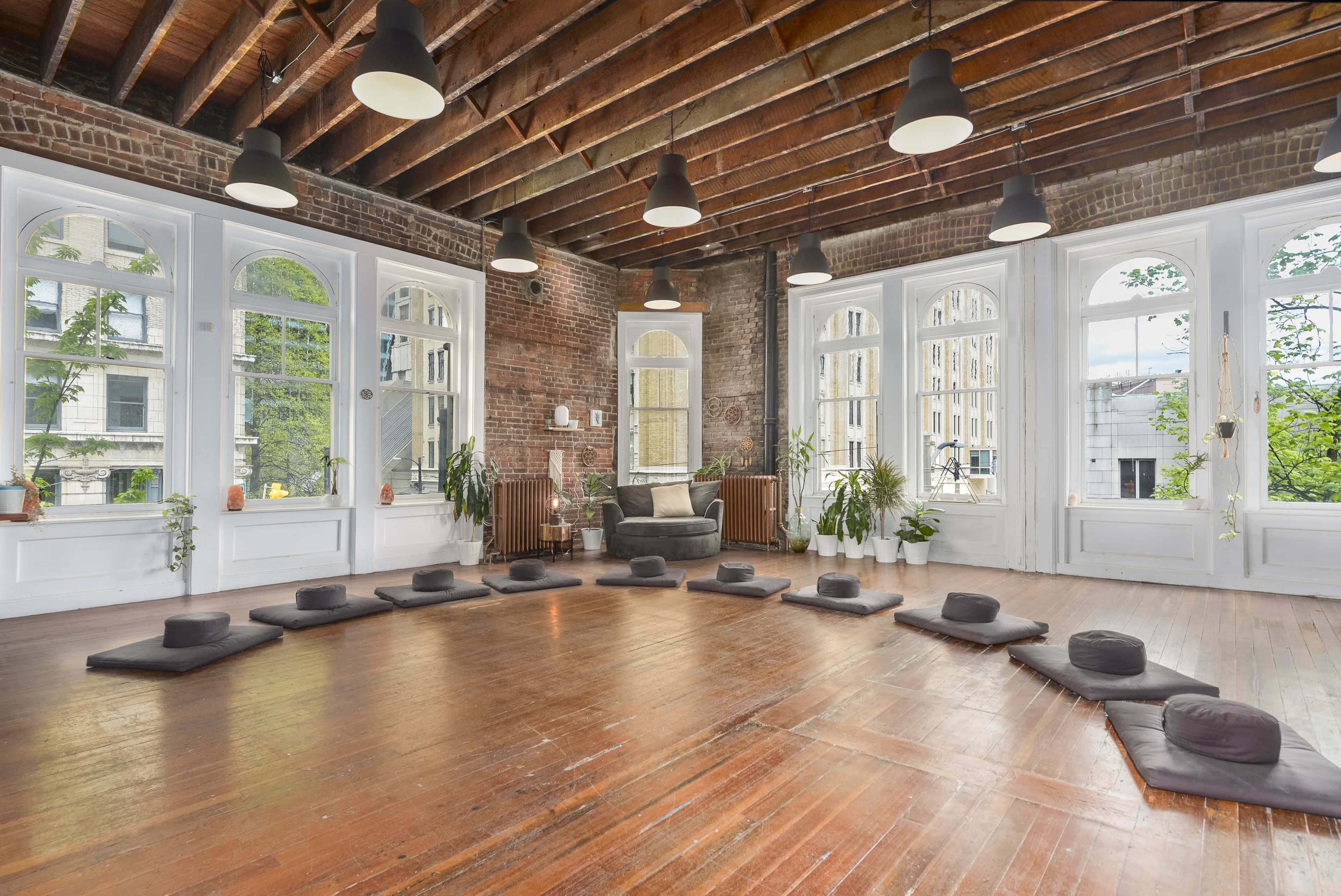 The room features a spacious layout with hardwood floors, plants along the windows, and seating arranged with cushions in a circular formation.