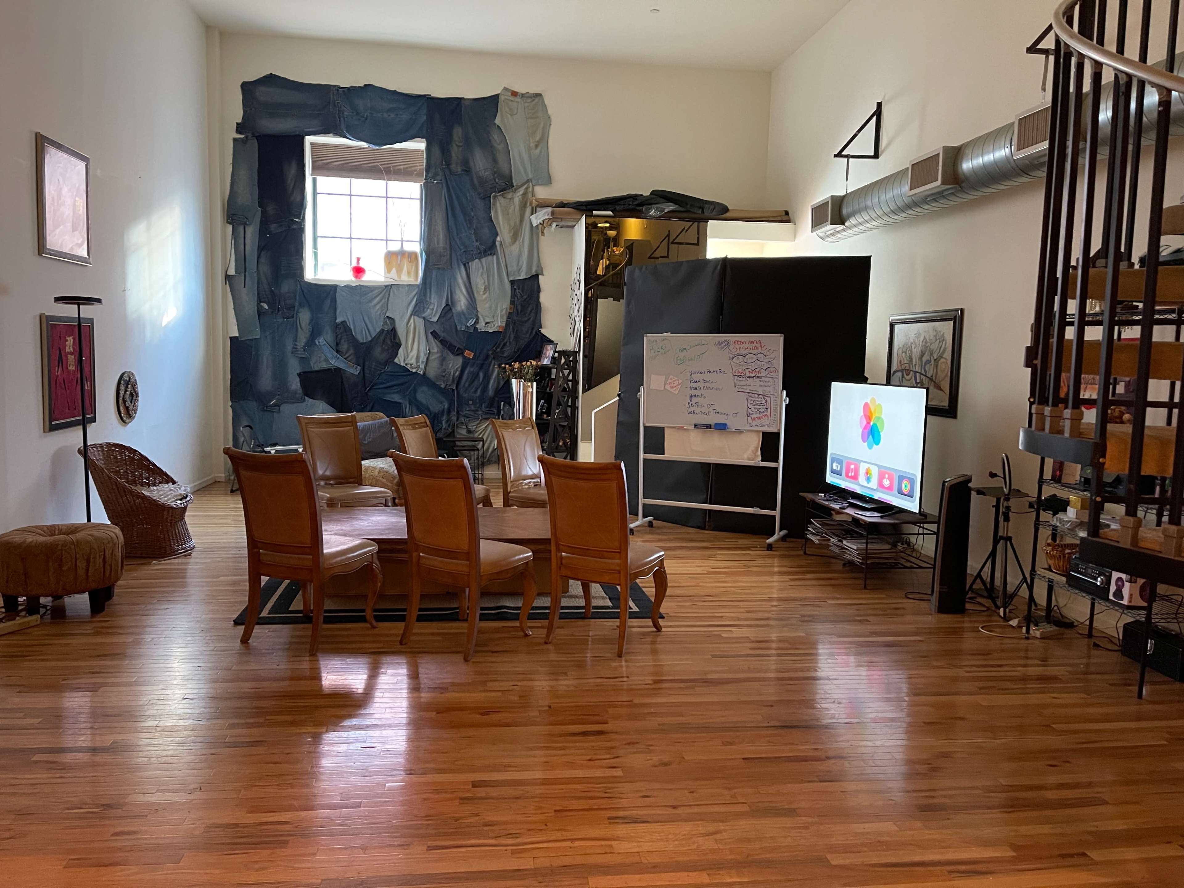 A spacious room features wooden flooring, a circular table surrounded by six chairs, a television, and a wall decorated with fabric.