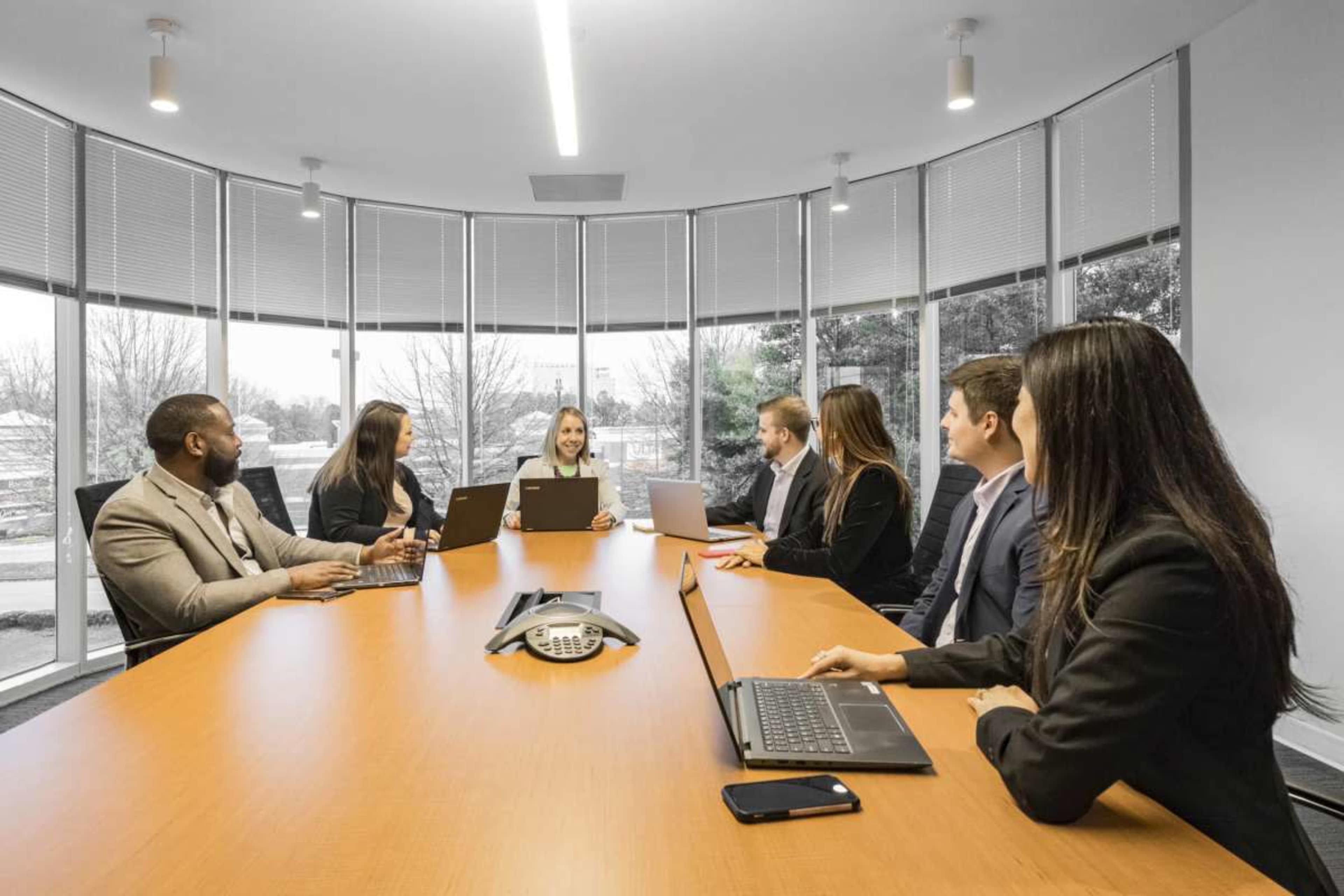 A group of seven professionals is seated around a large, oval conference table, engaged in a meeting with laptops open and a phone system in the center of the table.