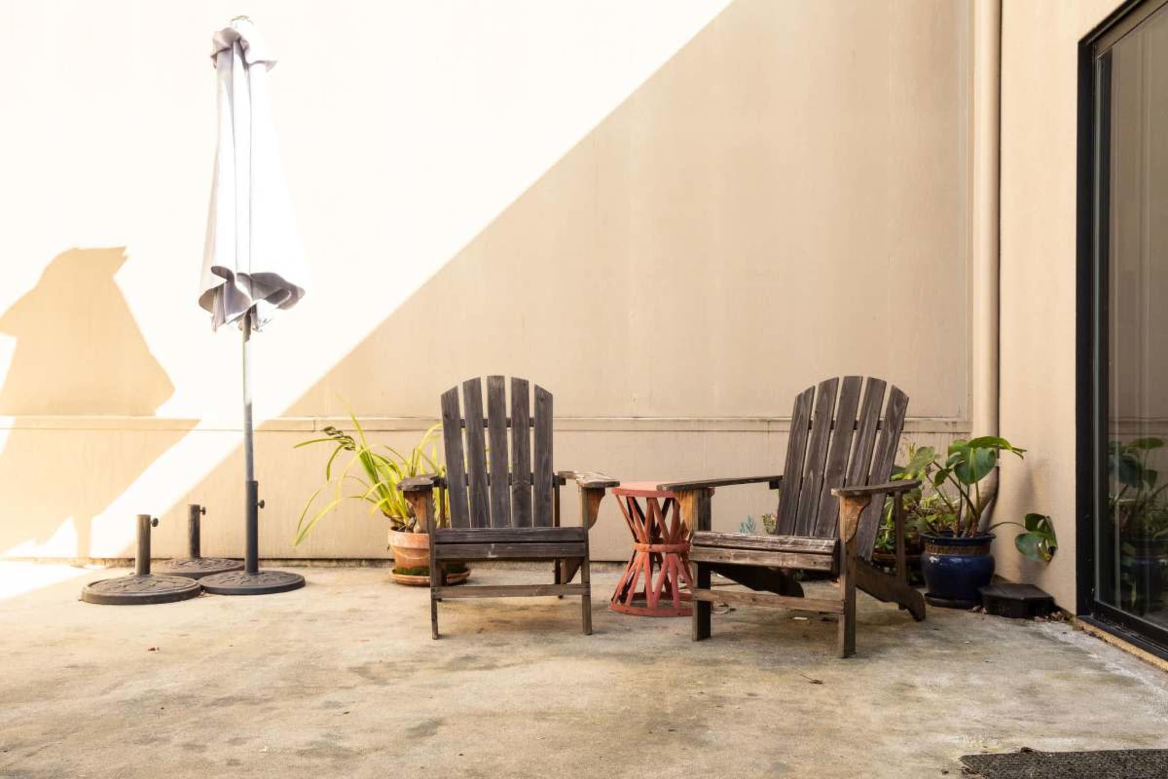 Two wooden Adirondack chairs and a small side table are placed on a concrete patio, accompanied by potted plants and a large umbrella stand.