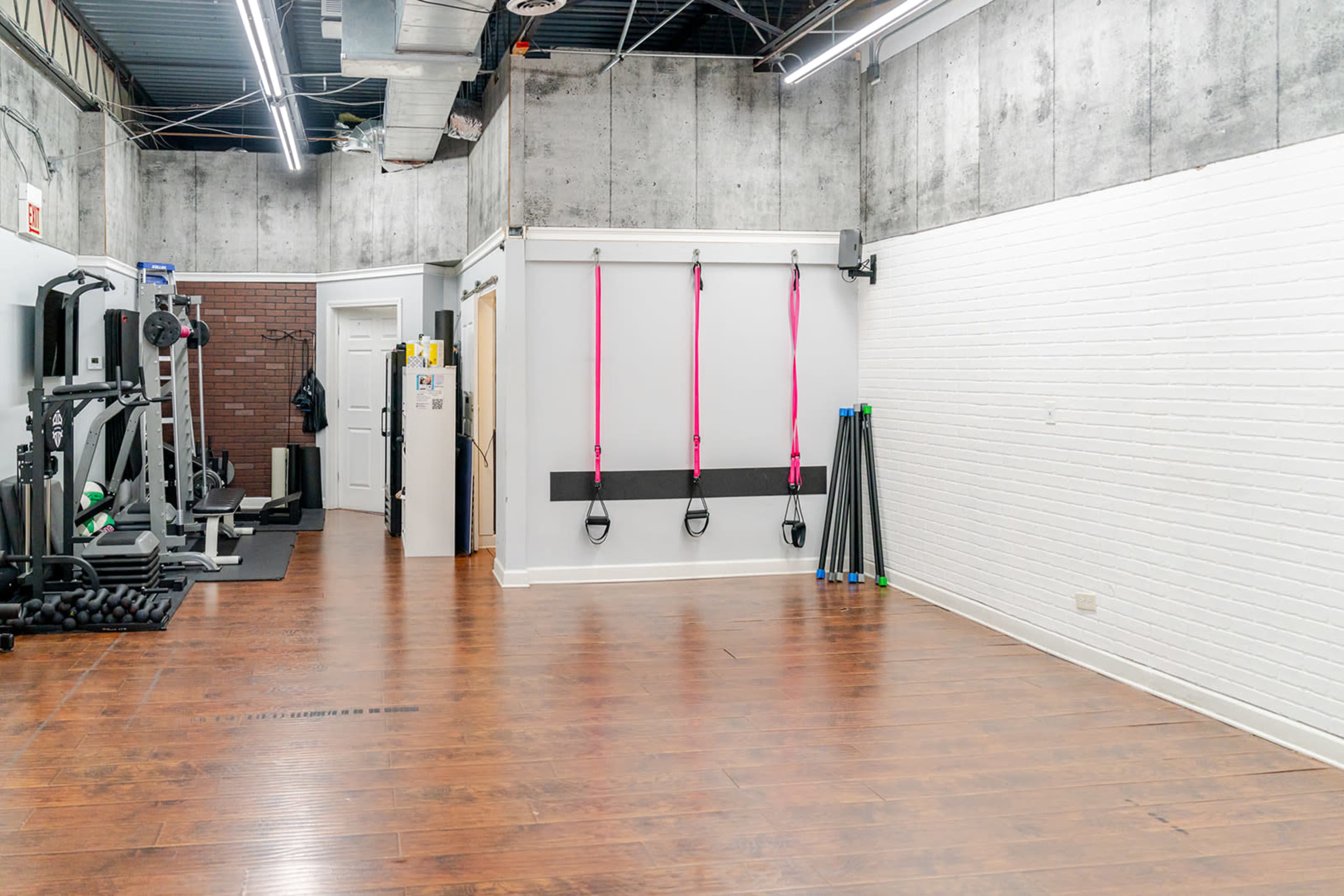 The image shows a spacious, minimally furnished gym with wooden flooring, gym equipment on one side, and colorful resistance bands hanging on the wall.