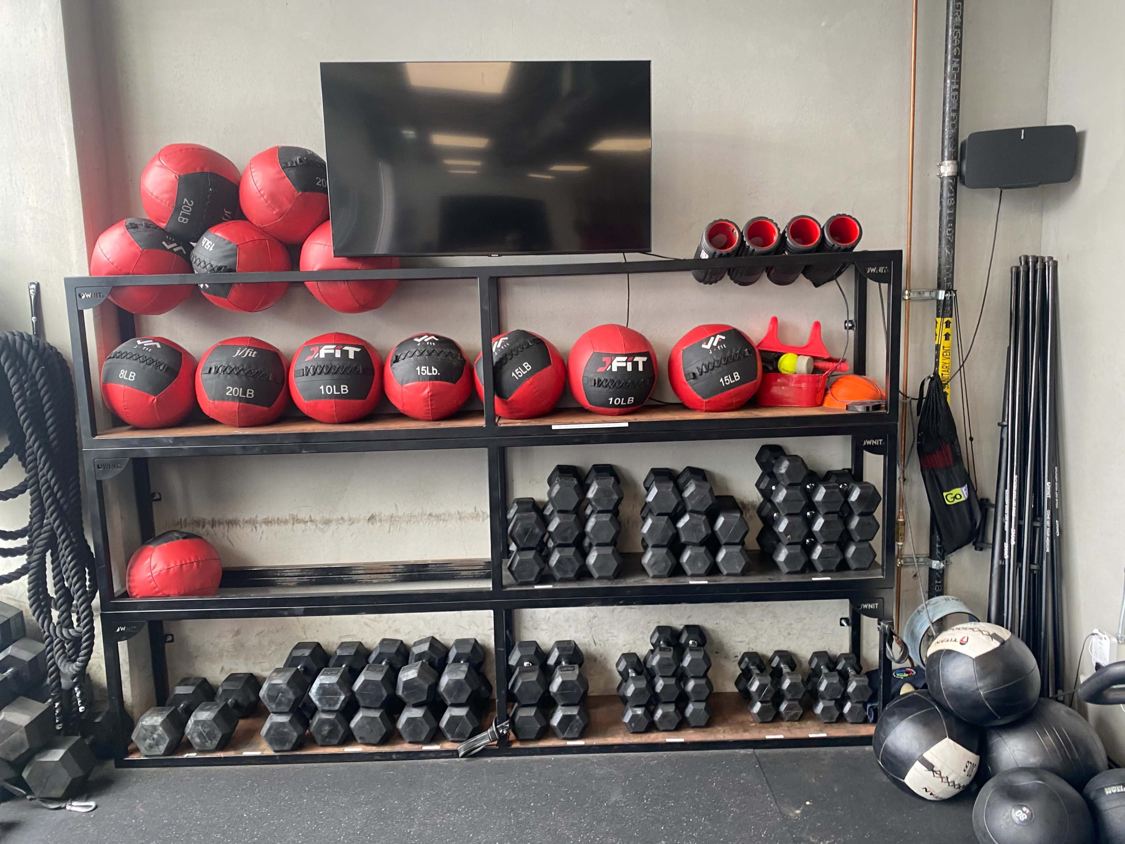 The image shows a well-organized gym storage rack containing various weights, including medicine balls and dumbbells, along with a television mounted on the wall.