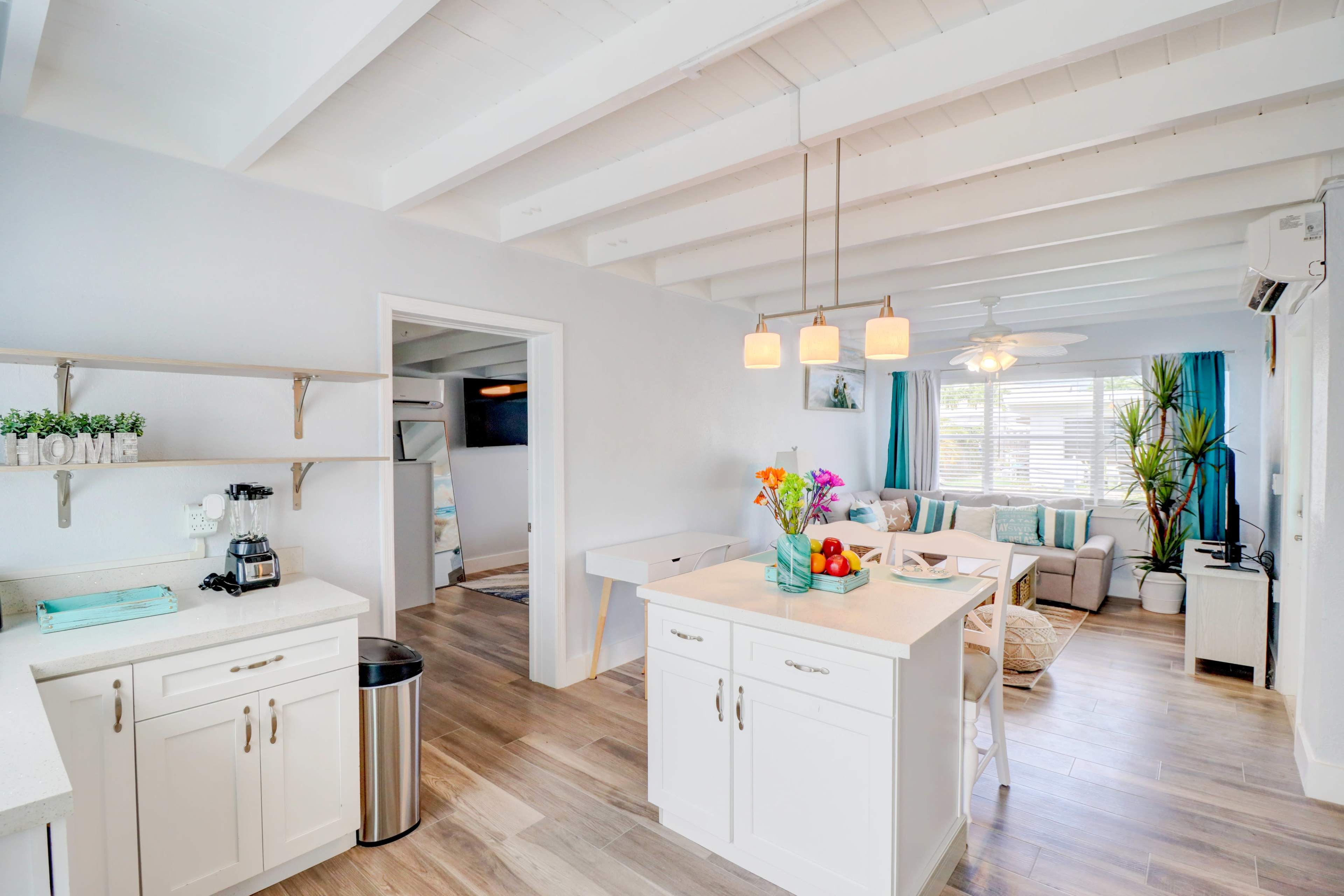 A modern kitchen area with white cabinets, a central island, and a living room featuring light furniture and large windows.