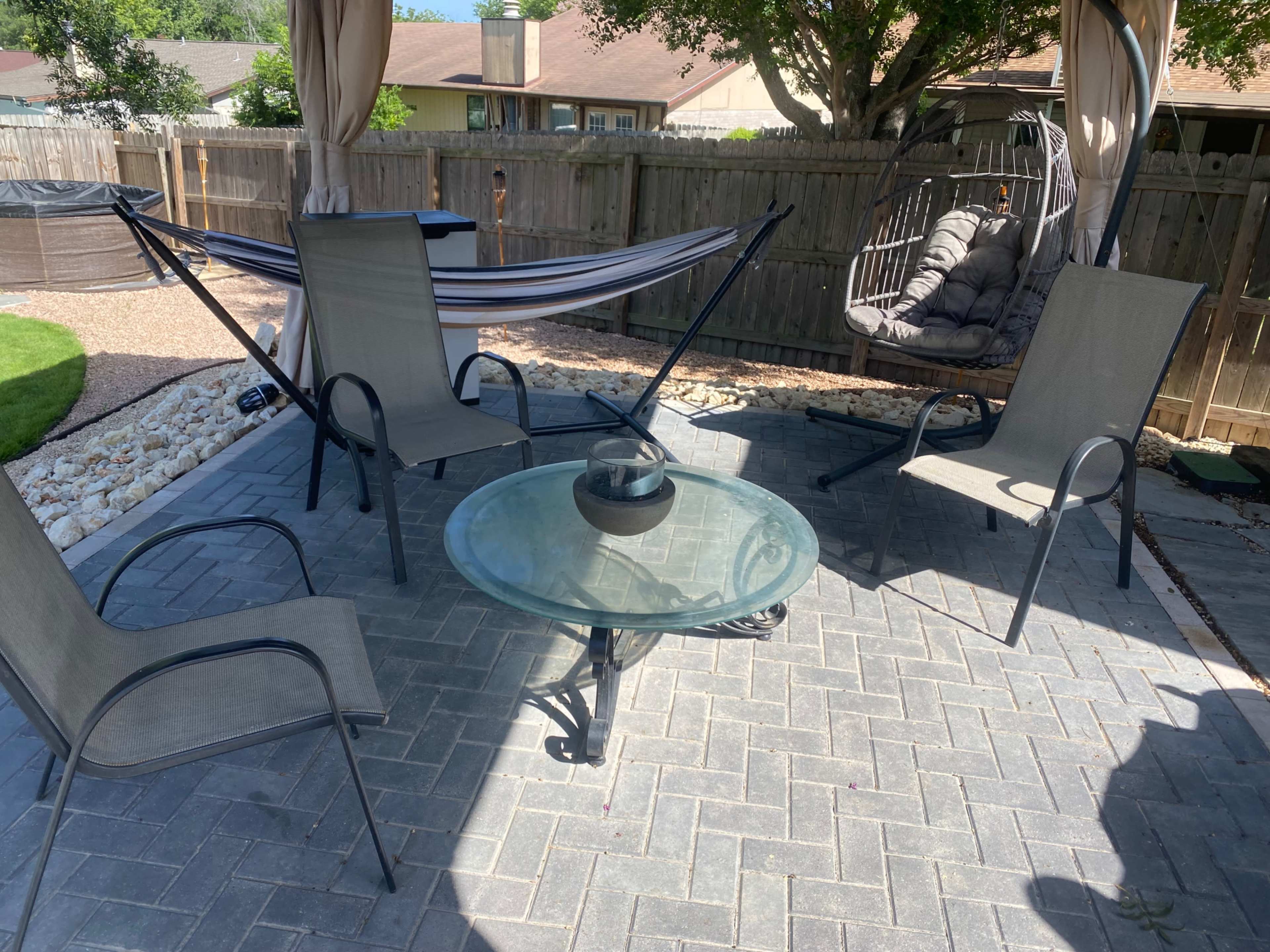 The image shows a patio area with a glass table surrounded by four chairs, a hammock in the background, and a hanging chair nearby.
