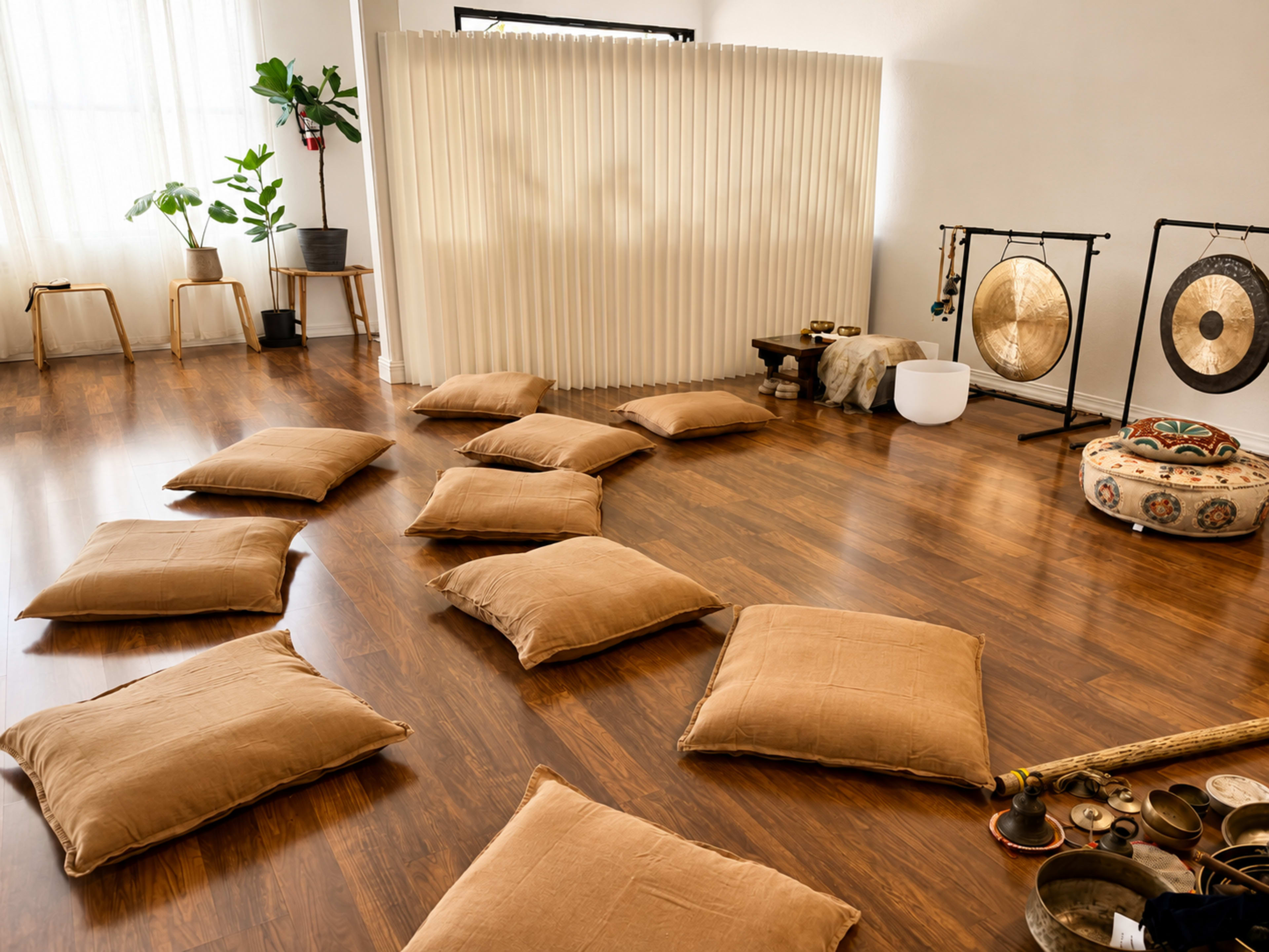 The image shows a spacious, wooden-floored room arranged with several large, tan cushions on the floor, a folding screen in the background, and various musical instruments, including gongs, placed nearby.