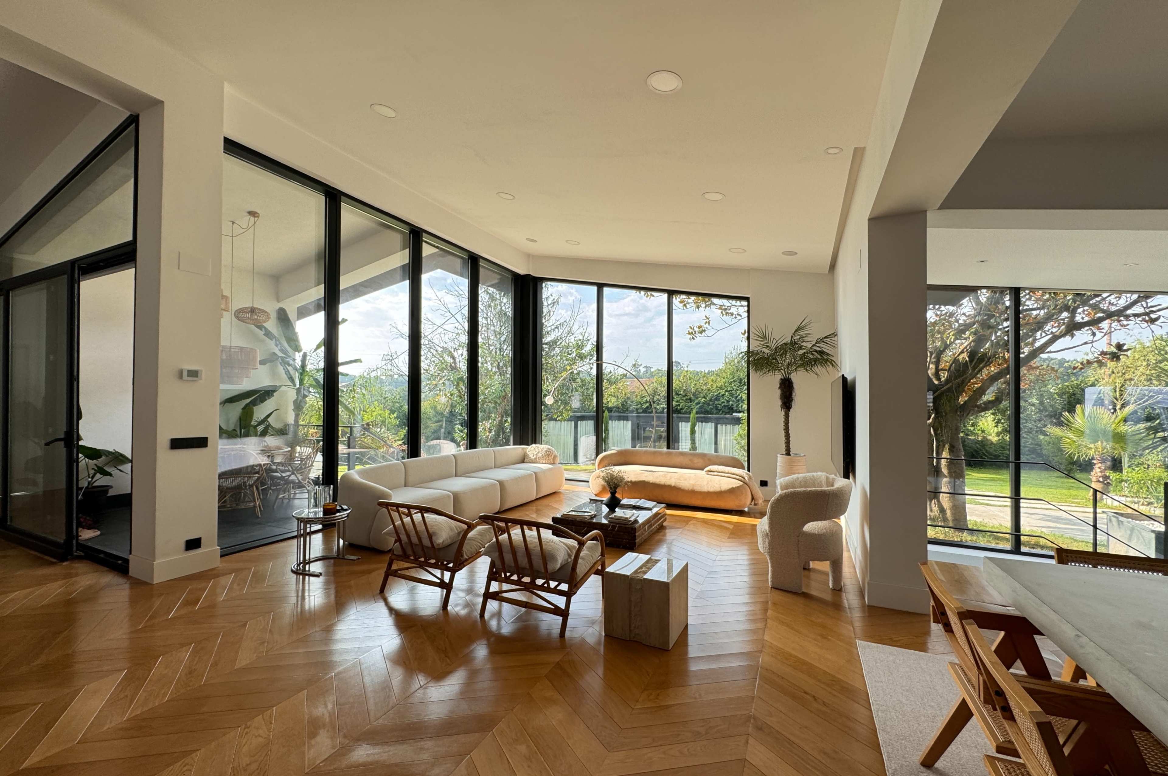 The image shows a modern living room with large glass windows, a light-colored sofa, and wooden furniture, overlooking a green outdoor space.