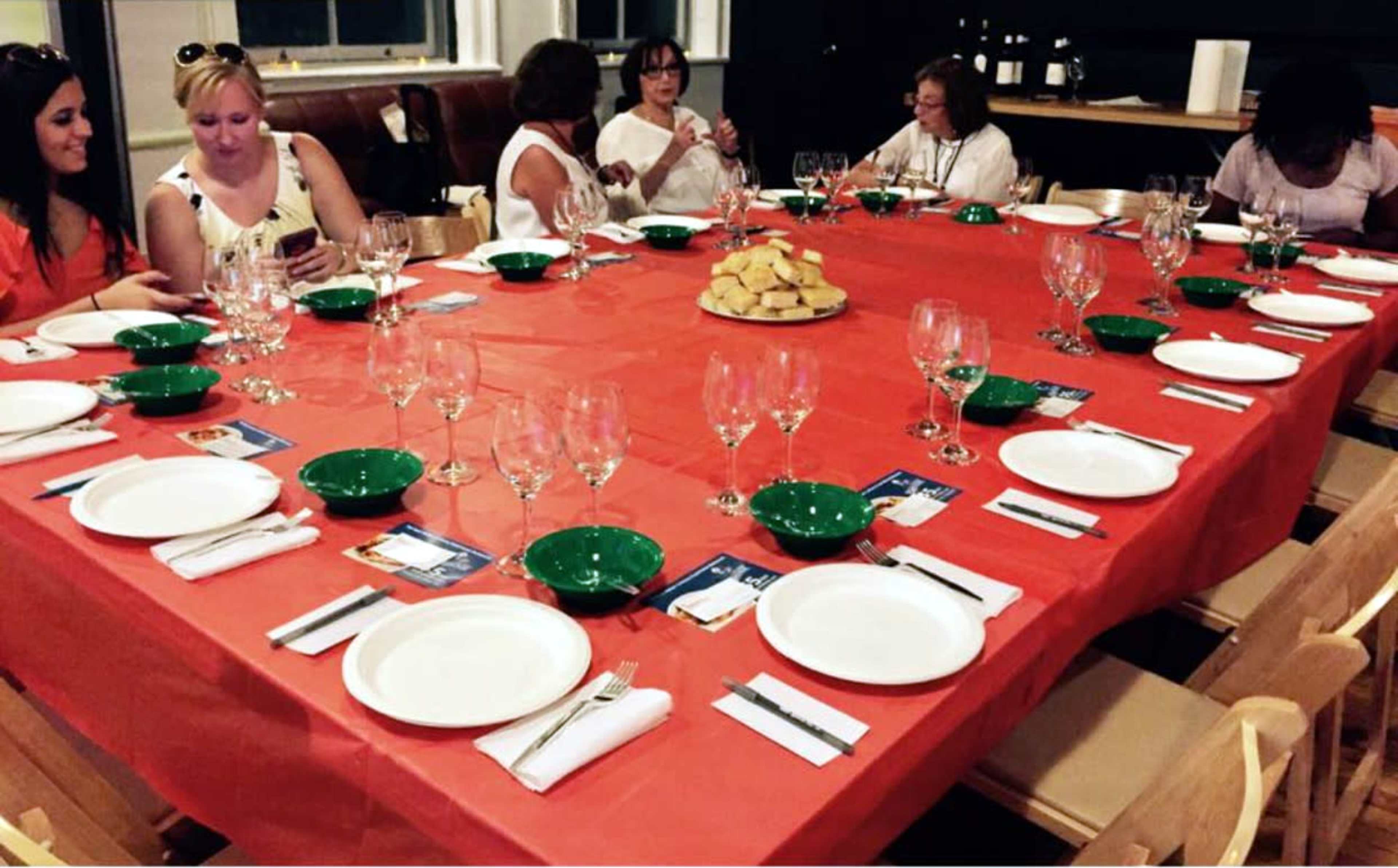 A long red table is set with white plates, green bowls, and empty wine glasses, while several people are seated and engaged in conversation.