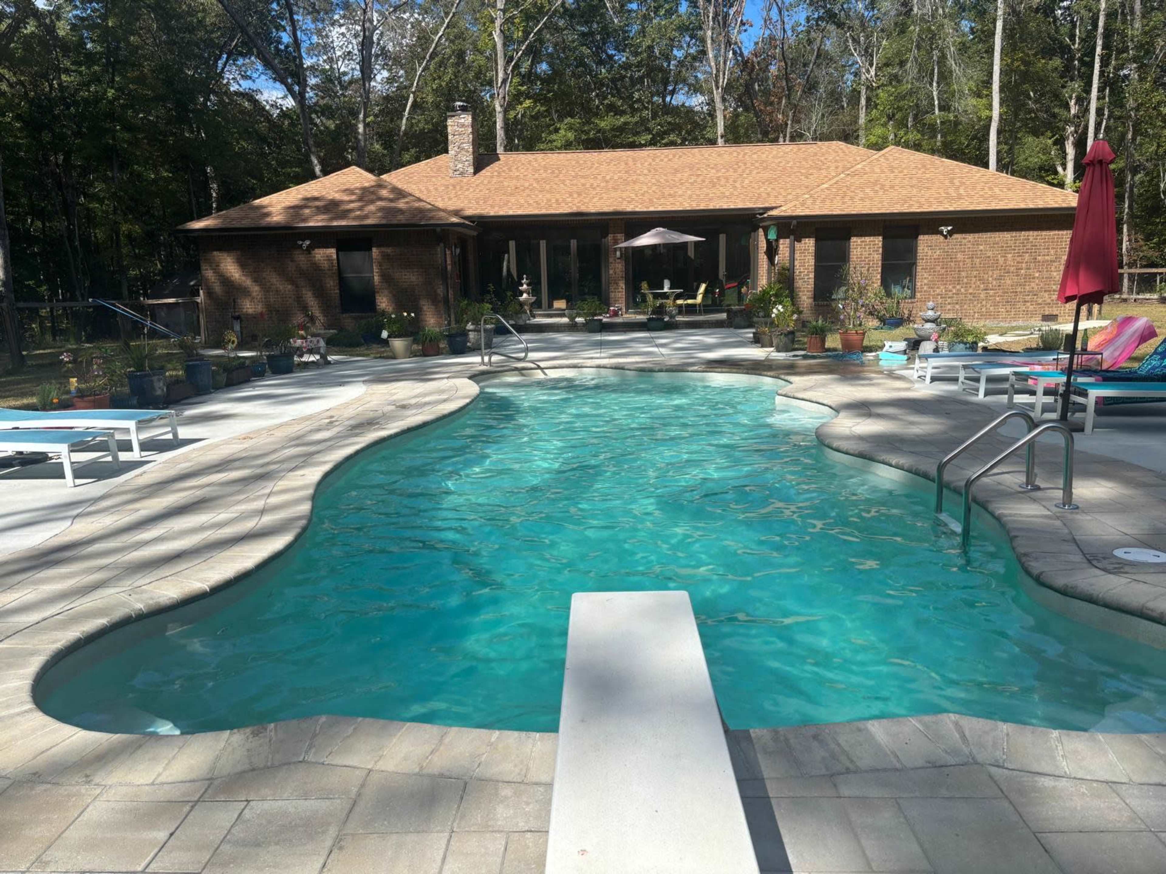 A swimming pool with a diving board is located in a backyard, adjacent to a brick house and surrounded by landscaped gardens.