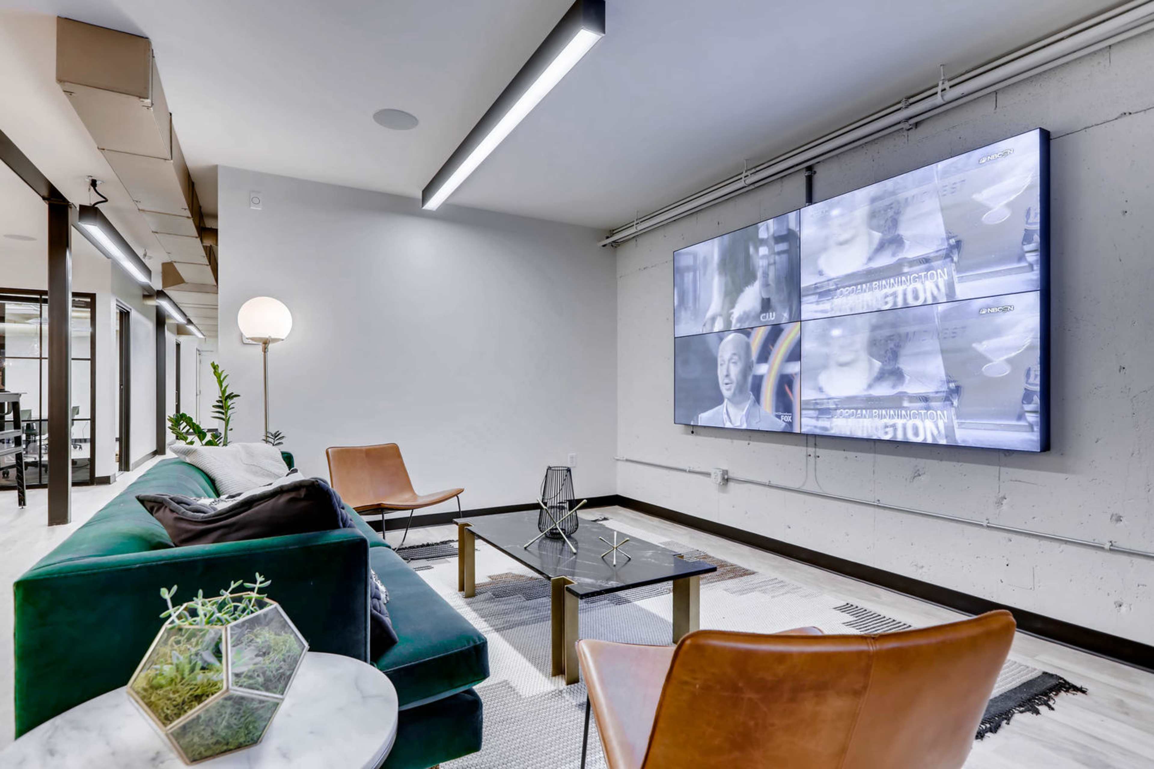 A modern lounge area features a large screen displaying video content, with a green sofa, two brown chairs, a glass table, and a floor lamp.