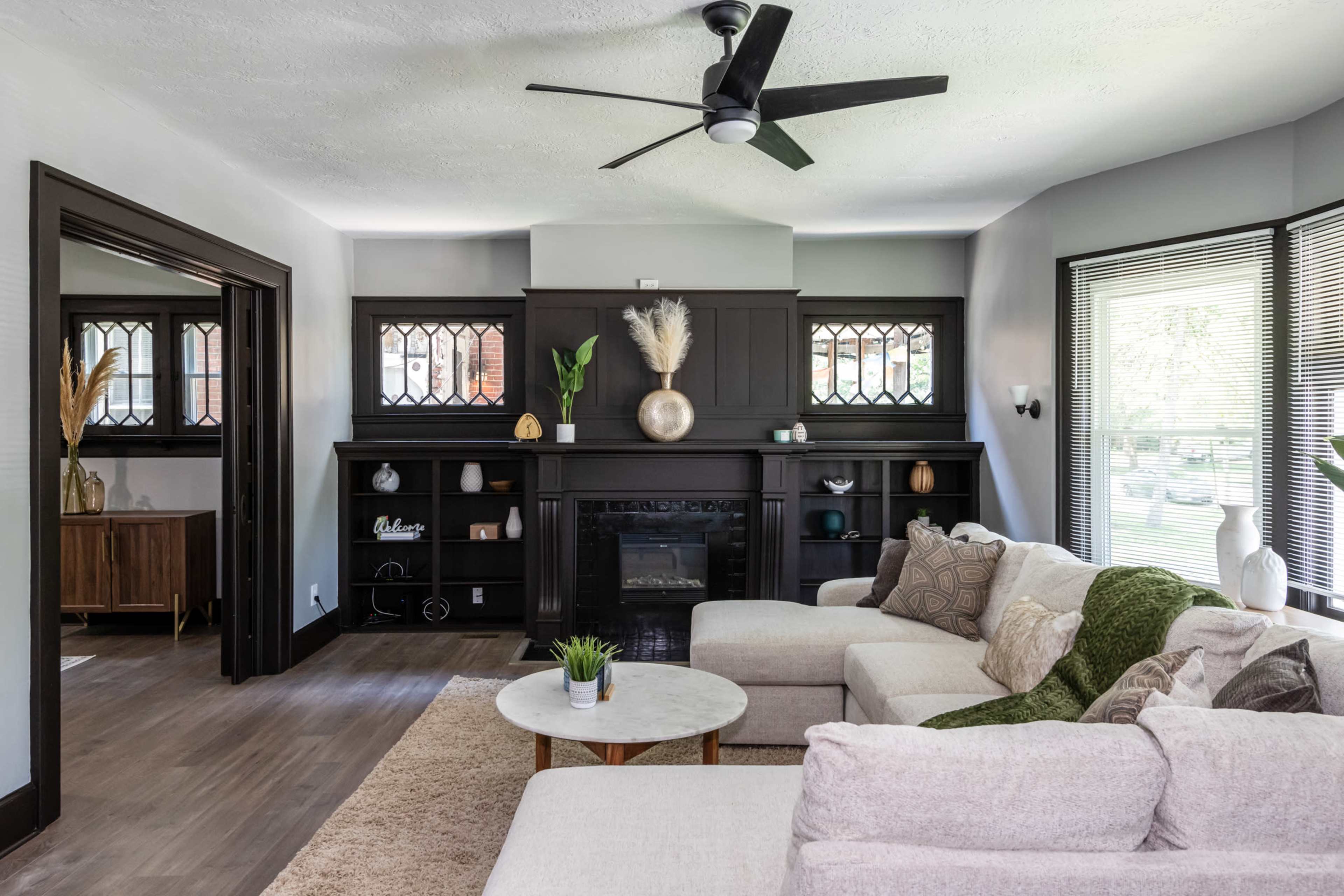 A modern living room with a light-colored sofa, a round coffee table, and a black fireplace surrounded by dark wood panels, accented by decorative items and plants.