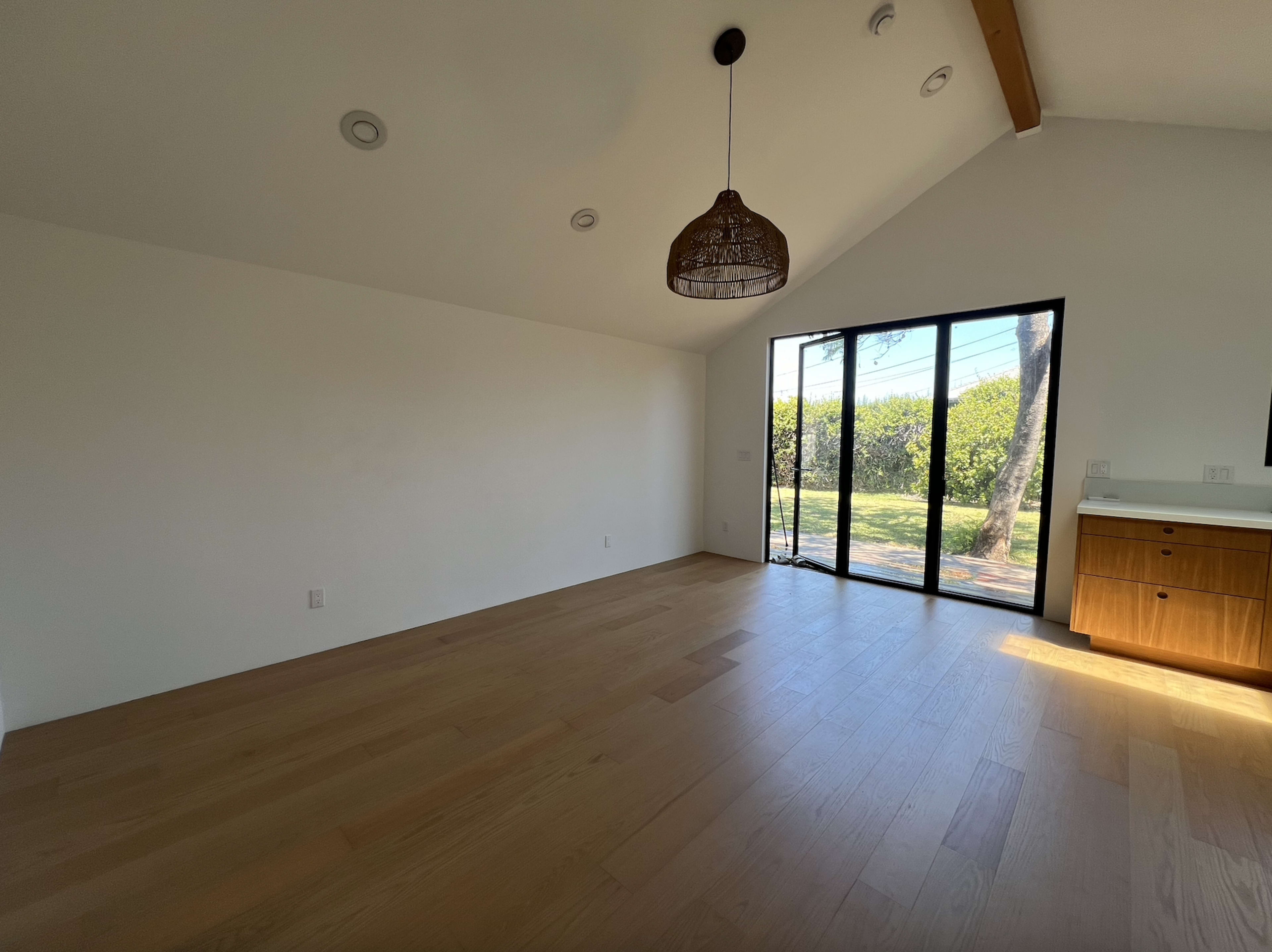 The image shows an empty room with wooden flooring, a high ceiling, and large glass doors leading to an outdoor area.