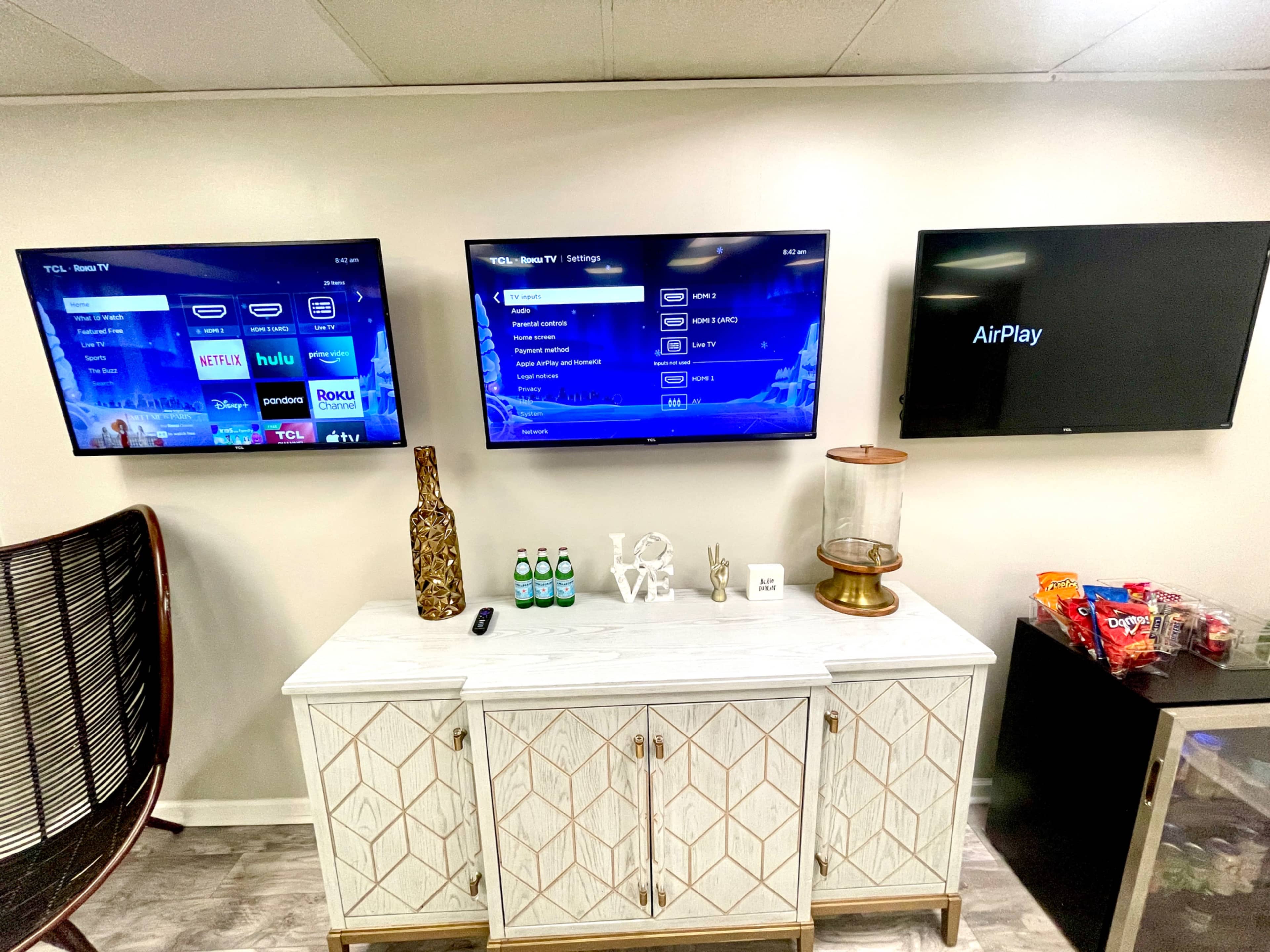 A modern room with three wall-mounted televisions displaying various settings, accompanied by a decorative cabinet featuring a vase, drinks, and snacks.