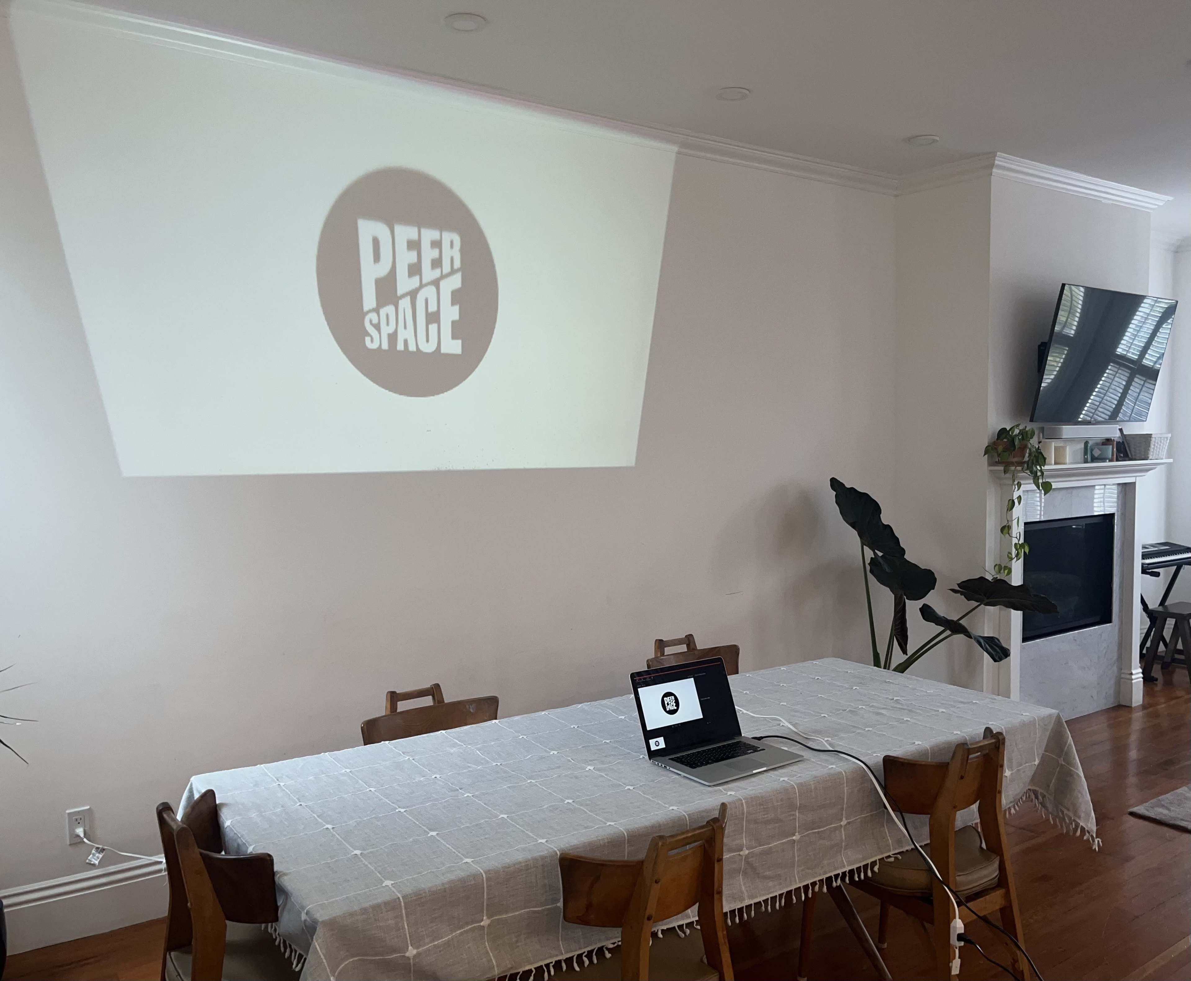 A laptop is placed on a dining table in a room where the PeerSpace logo is projected onto the wall.