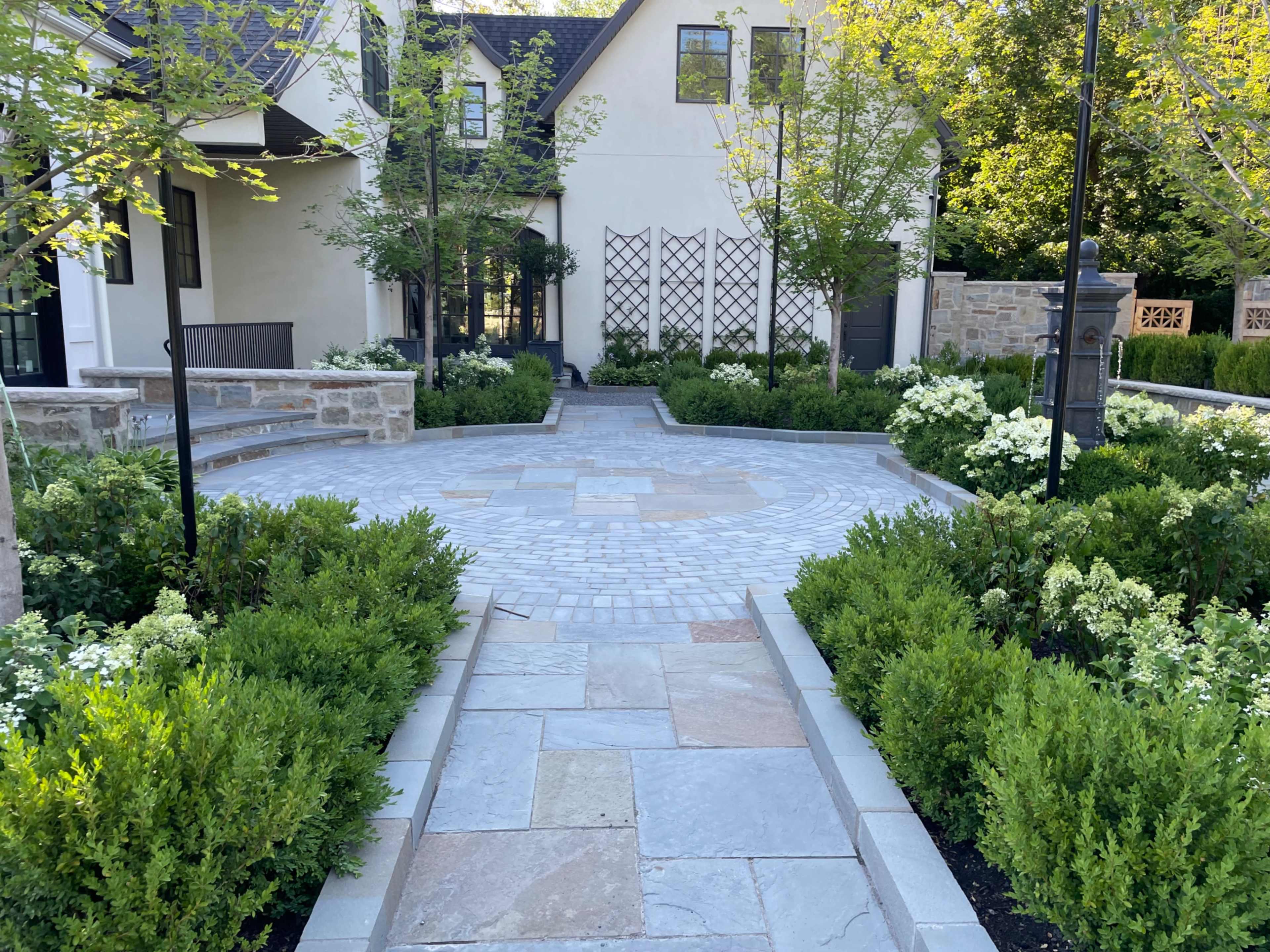 The image shows a landscaped garden walkway featuring stone pavers, surrounded by lush greenery and flowering plants, leading to a building.