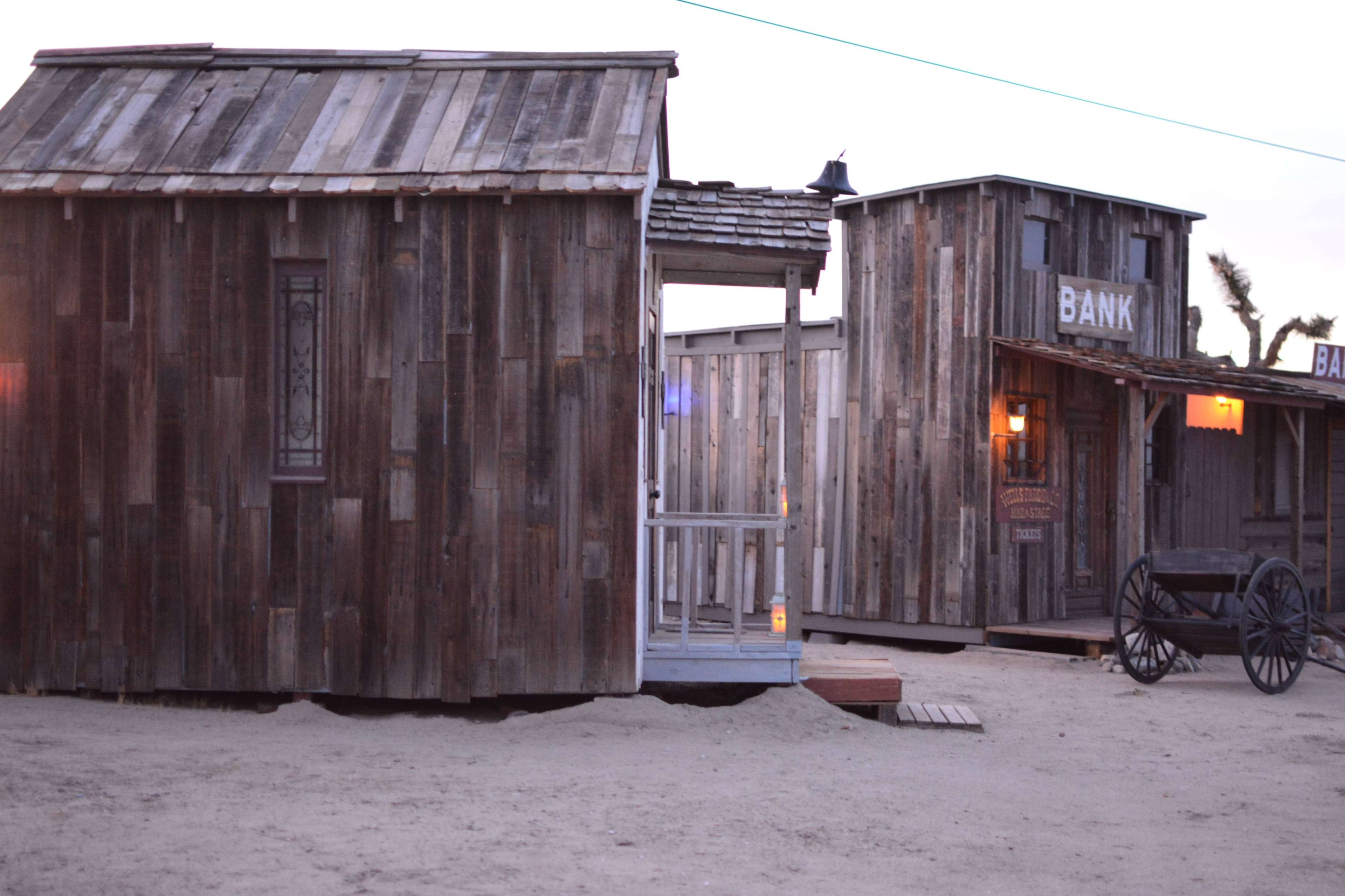 A western-style ghost town with wooden buildings, including a bank and a small house, set against a sandy landscape.