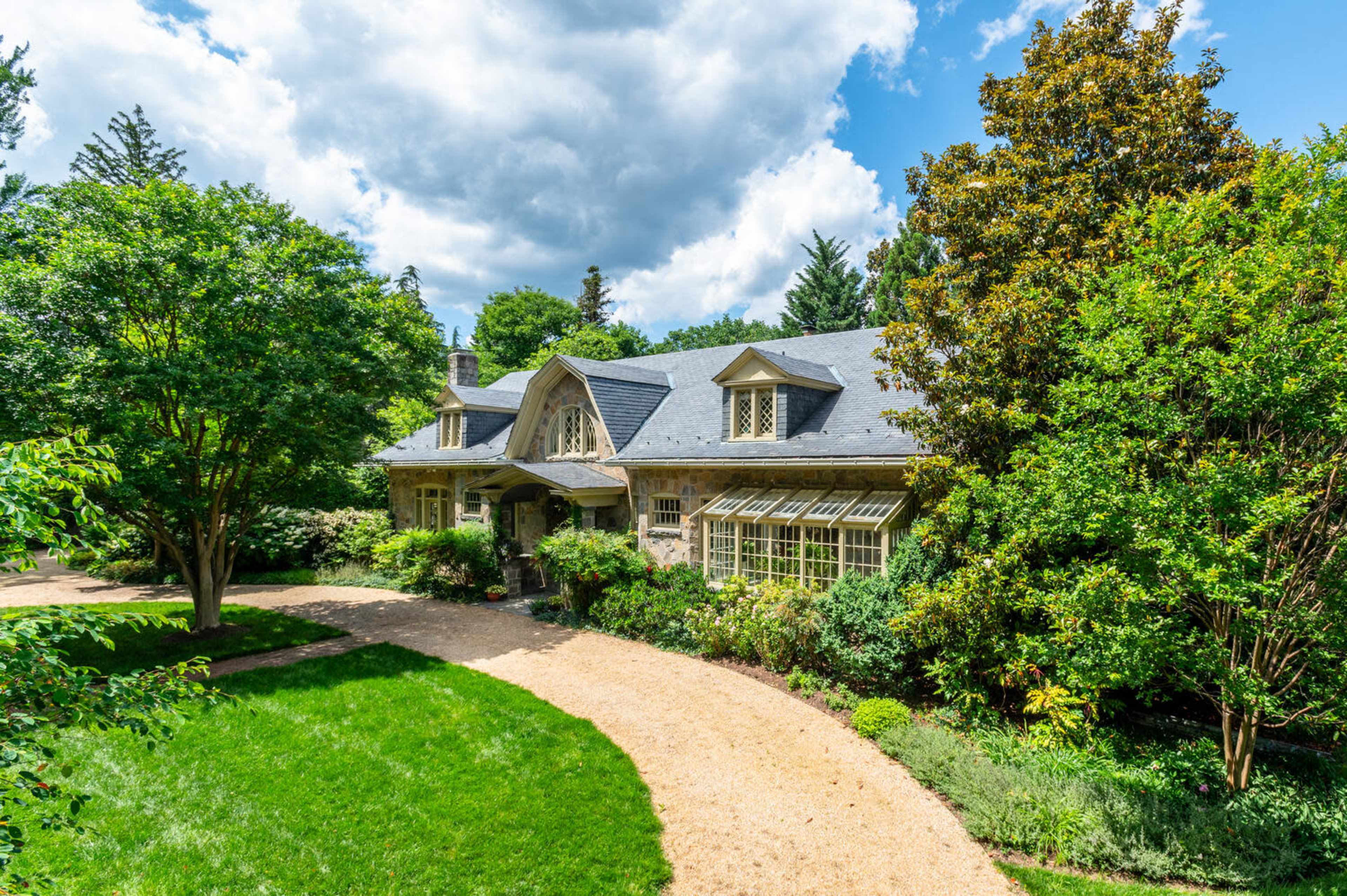 A large, stone-faced house with a gabled roof is set among lush greenery and a winding gravel driveway.