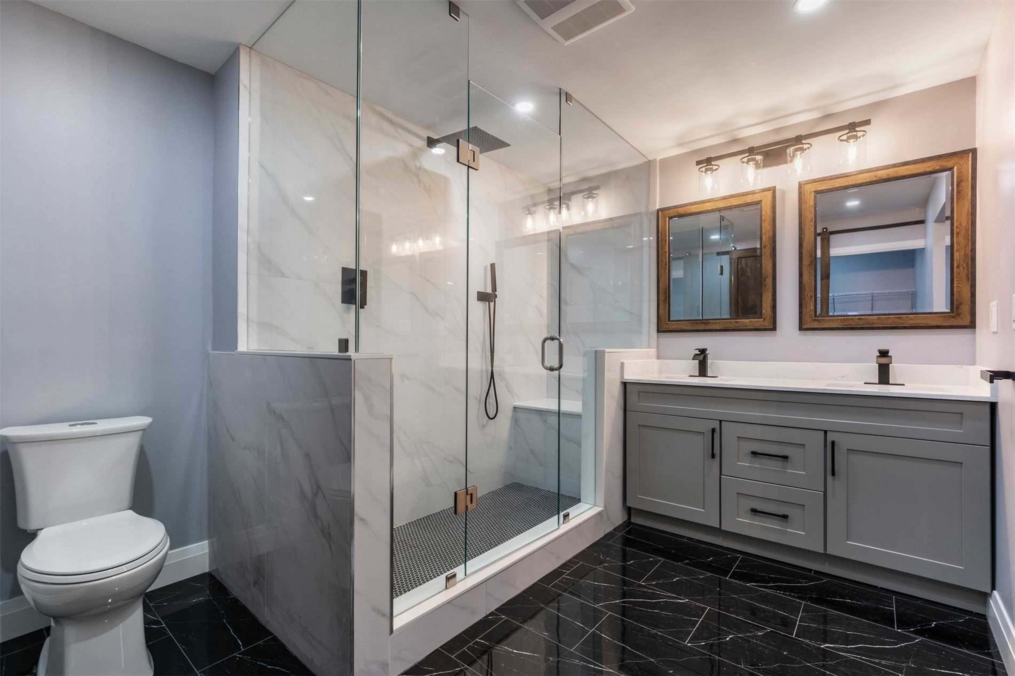 The image shows a modern bathroom featuring a frameless glass shower, a double sink vanity with mirrors, and a toilet.