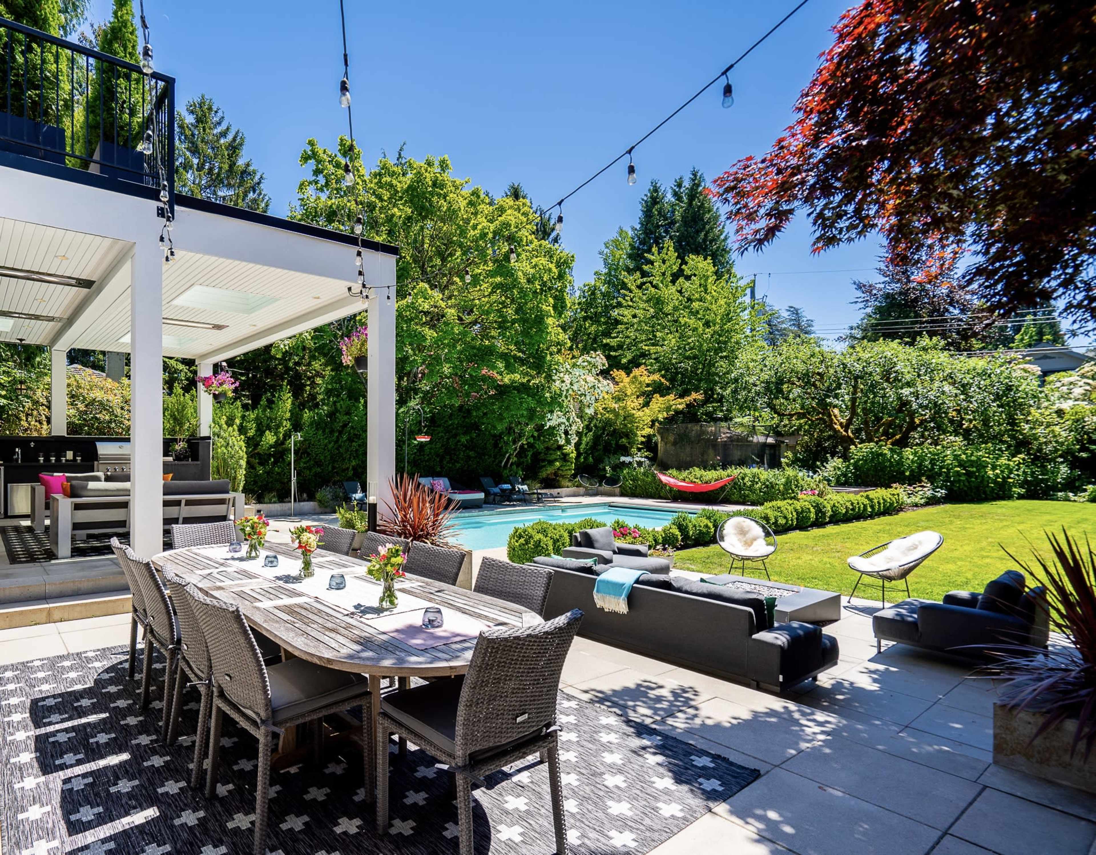 The image depicts a spacious outdoor dining area with a large table and chairs, surrounded by lush greenery and a swimming pool in the background.