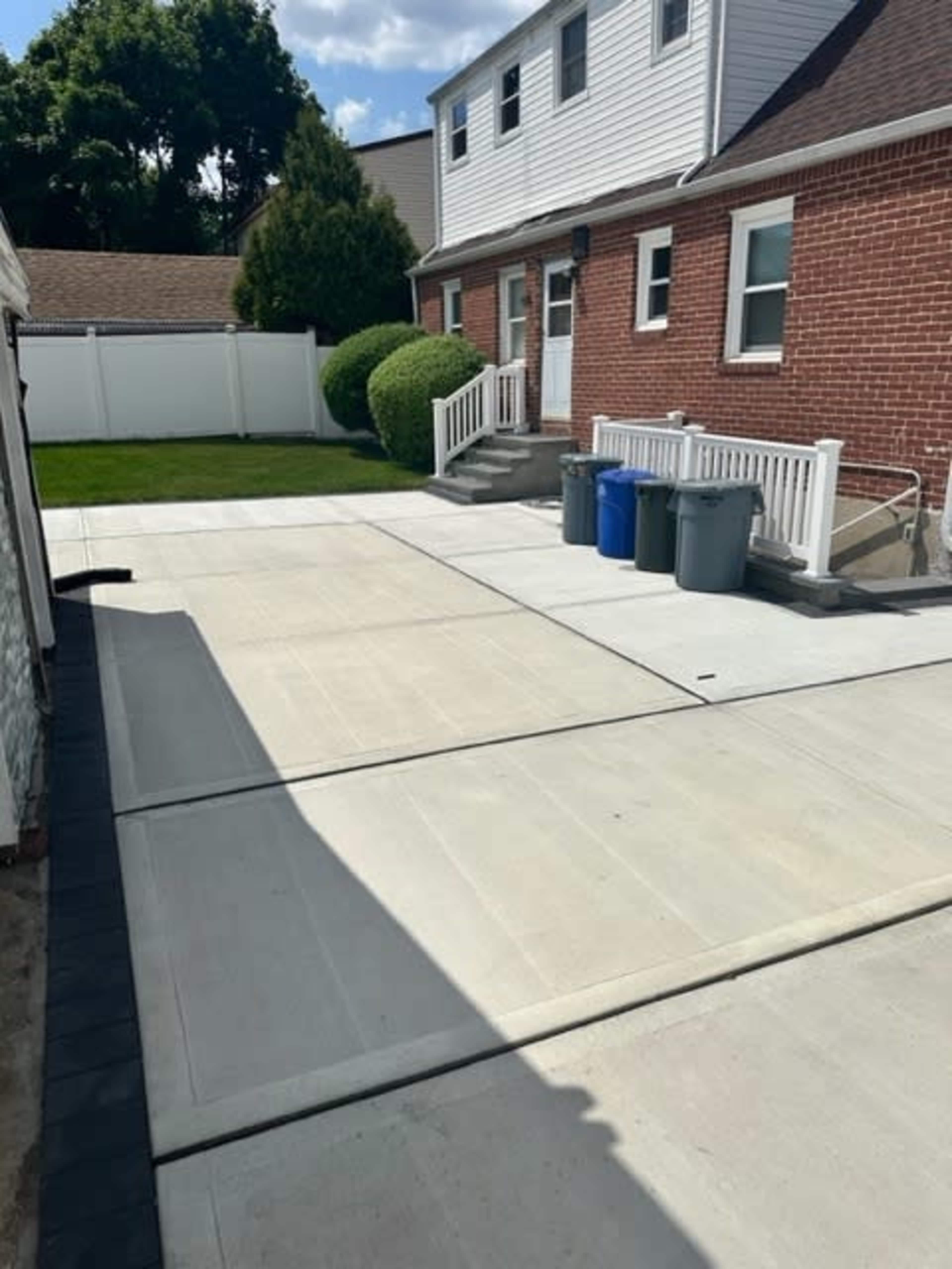 A driveway made of concrete, leading to a brick house, with blue and gray trash bins lined up on the side and a grassy yard behind a white fence.