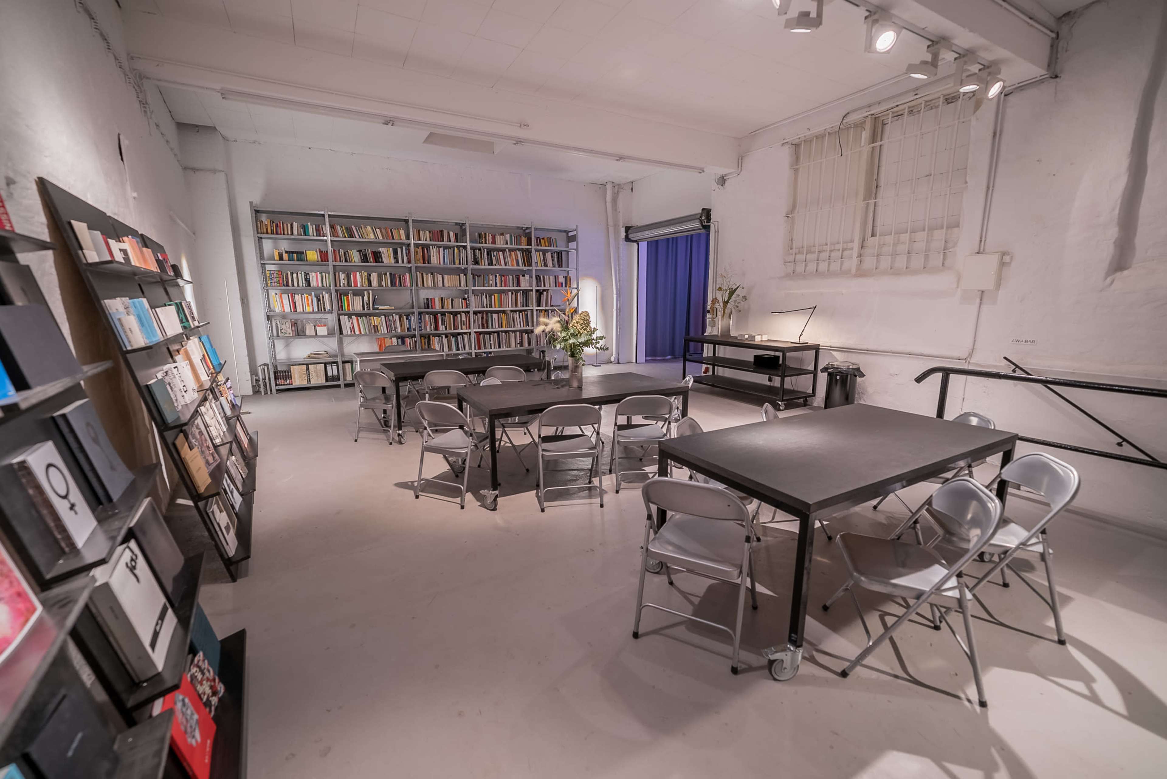 A bright, airy room features several black tables and chairs, a bookshelf filled with books, and a blue curtain draped against a wall.