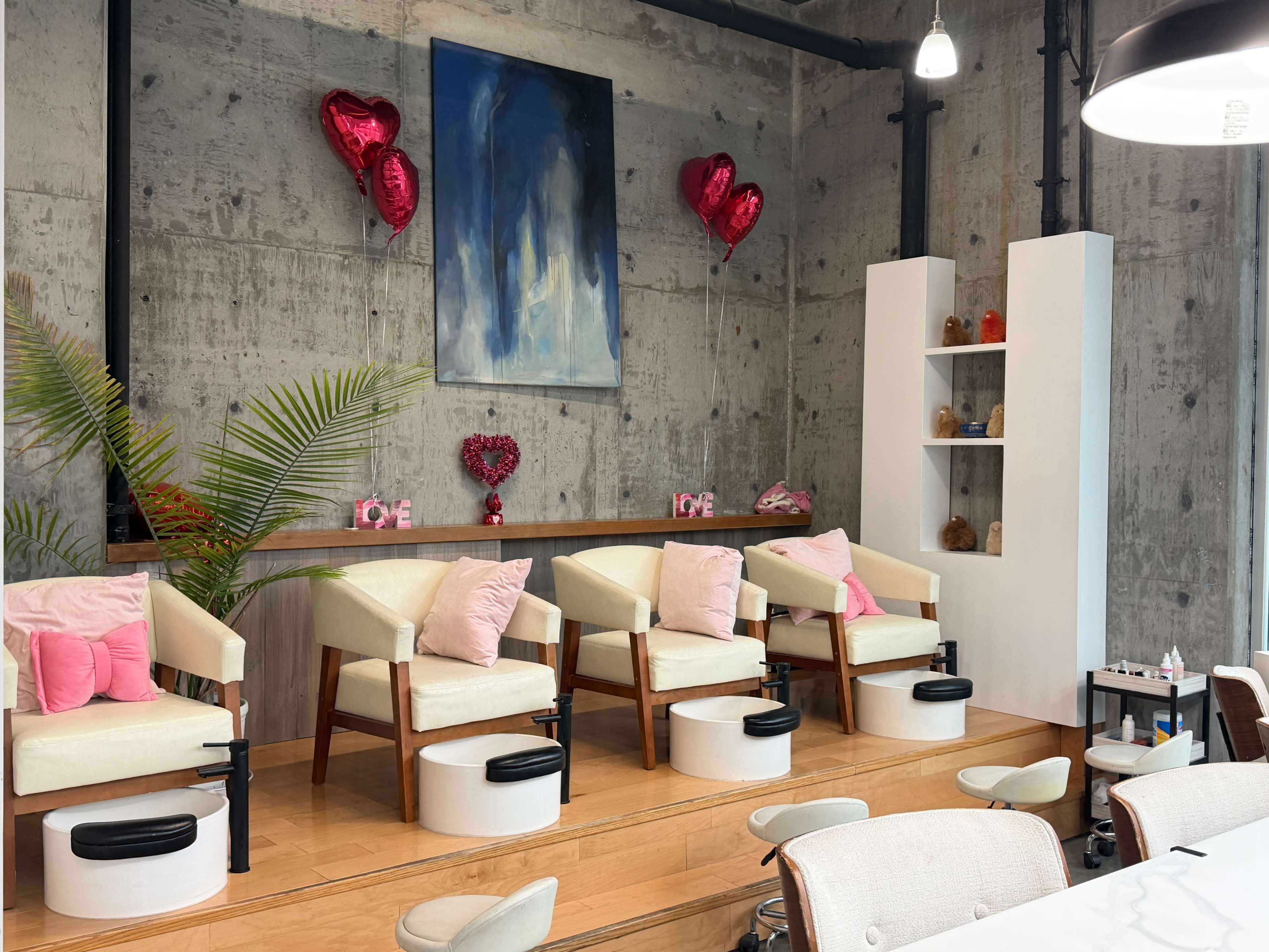 The image shows a modern nail salon with three pedicure chairs positioned on a wooden platform, decorated with pink hearts and balloons.