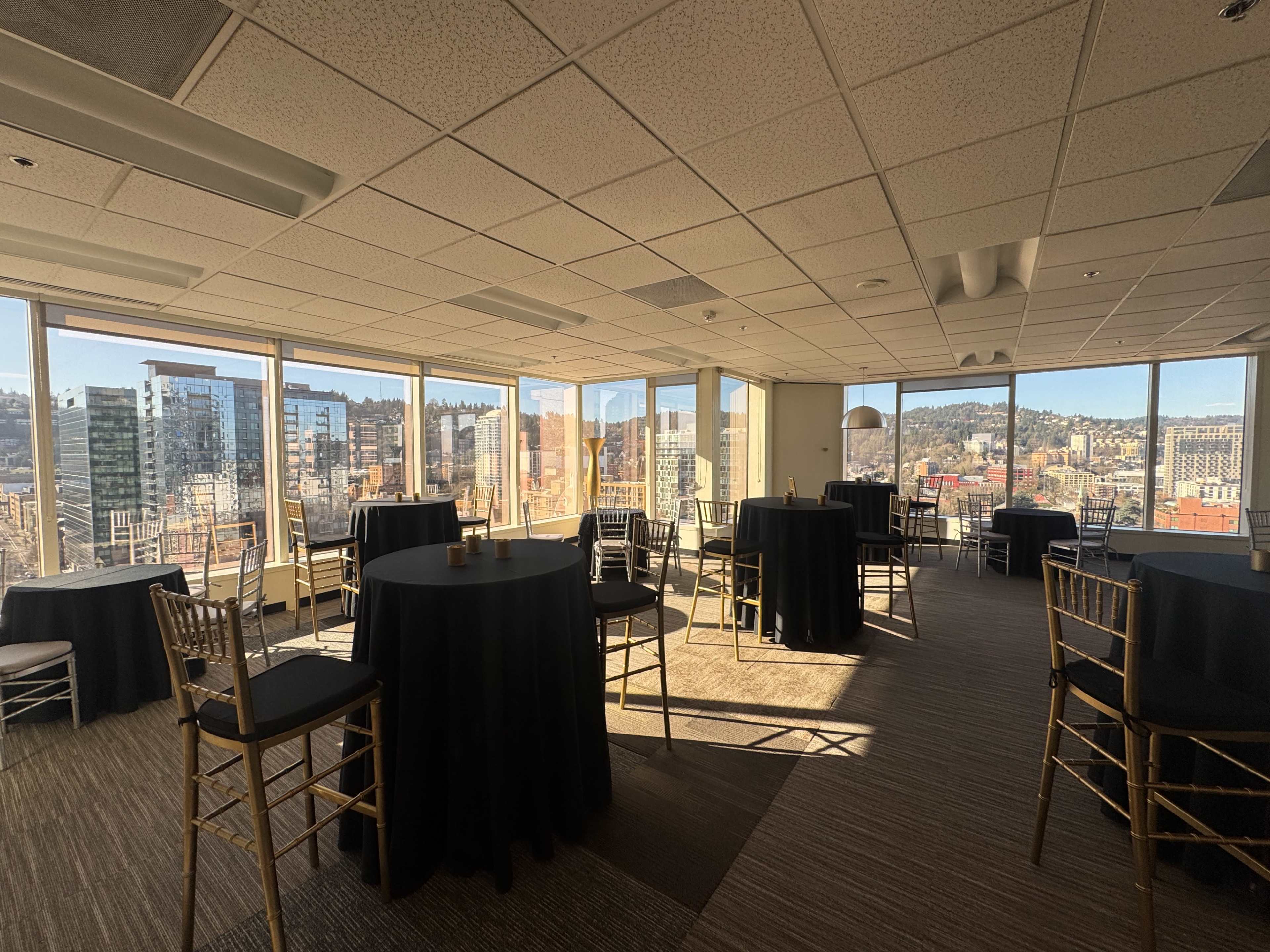 The image shows a large event space with several black-covered tables and large windows offering a view of the city skyline.