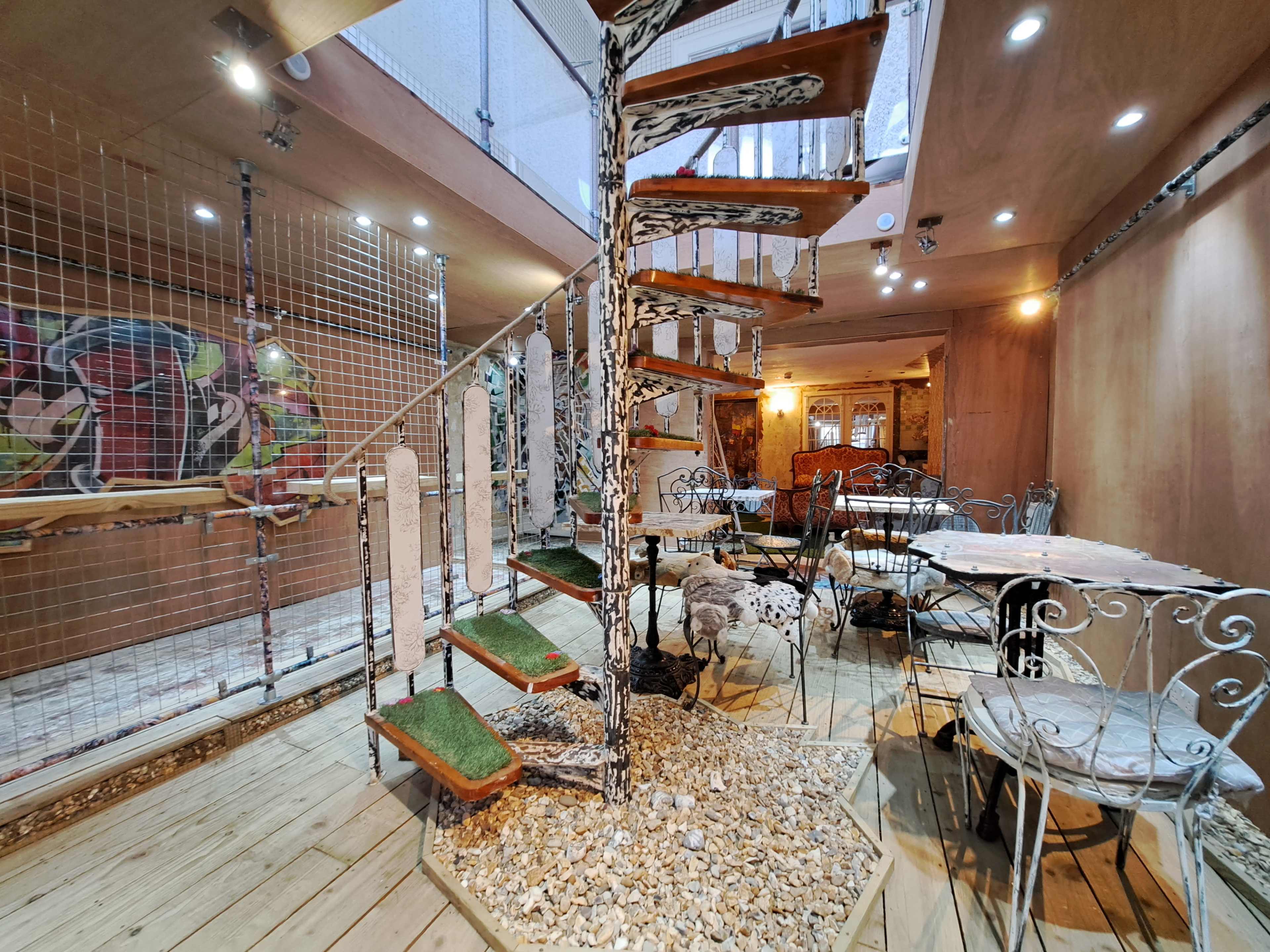 A modern indoor space with a spiral staircase, circular tables with wrought iron chairs, and decorative elements like a gravel area and wall art.