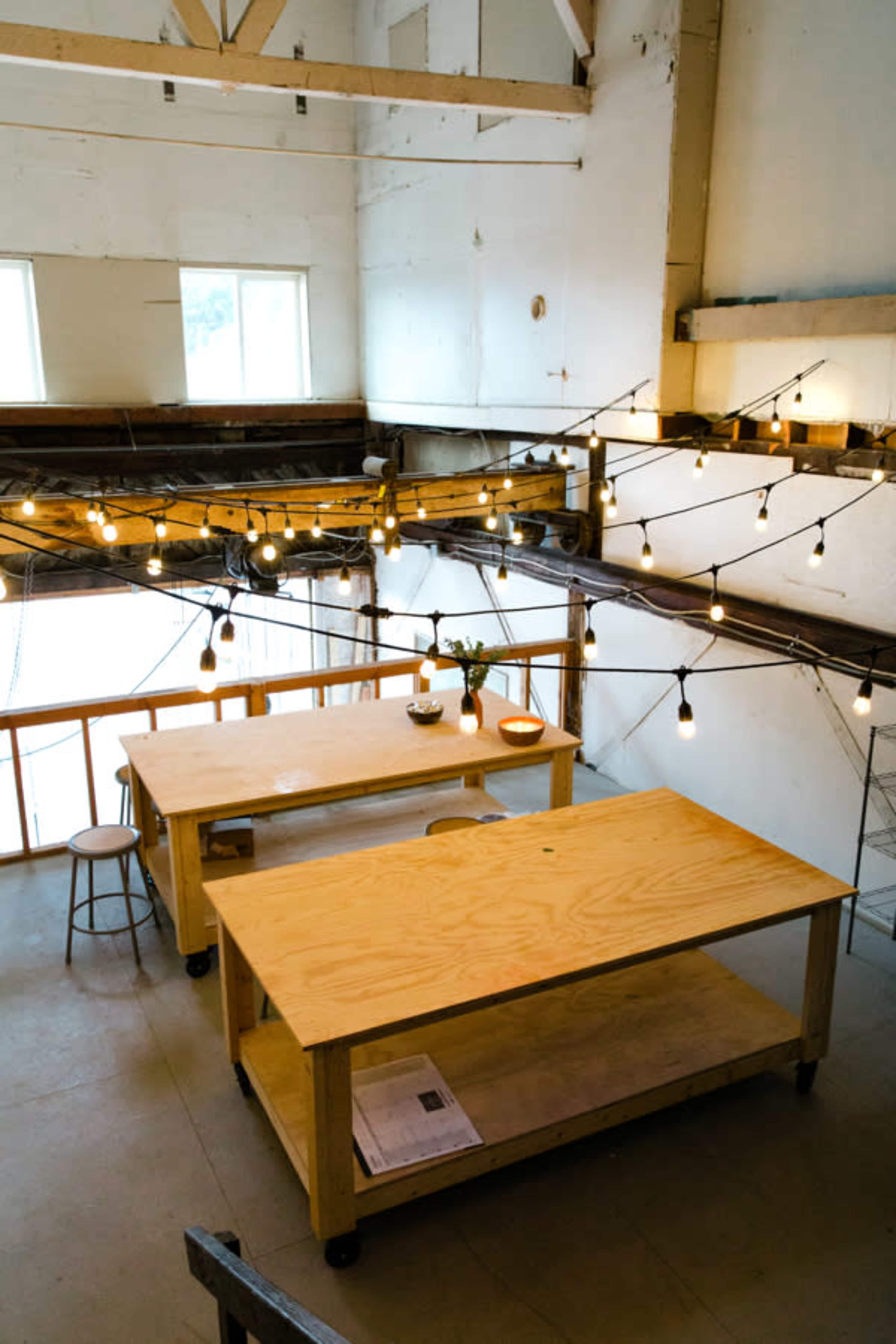 The image shows a spacious workshop with wooden tables, a stool, and string lights hanging above in a bright, open area.