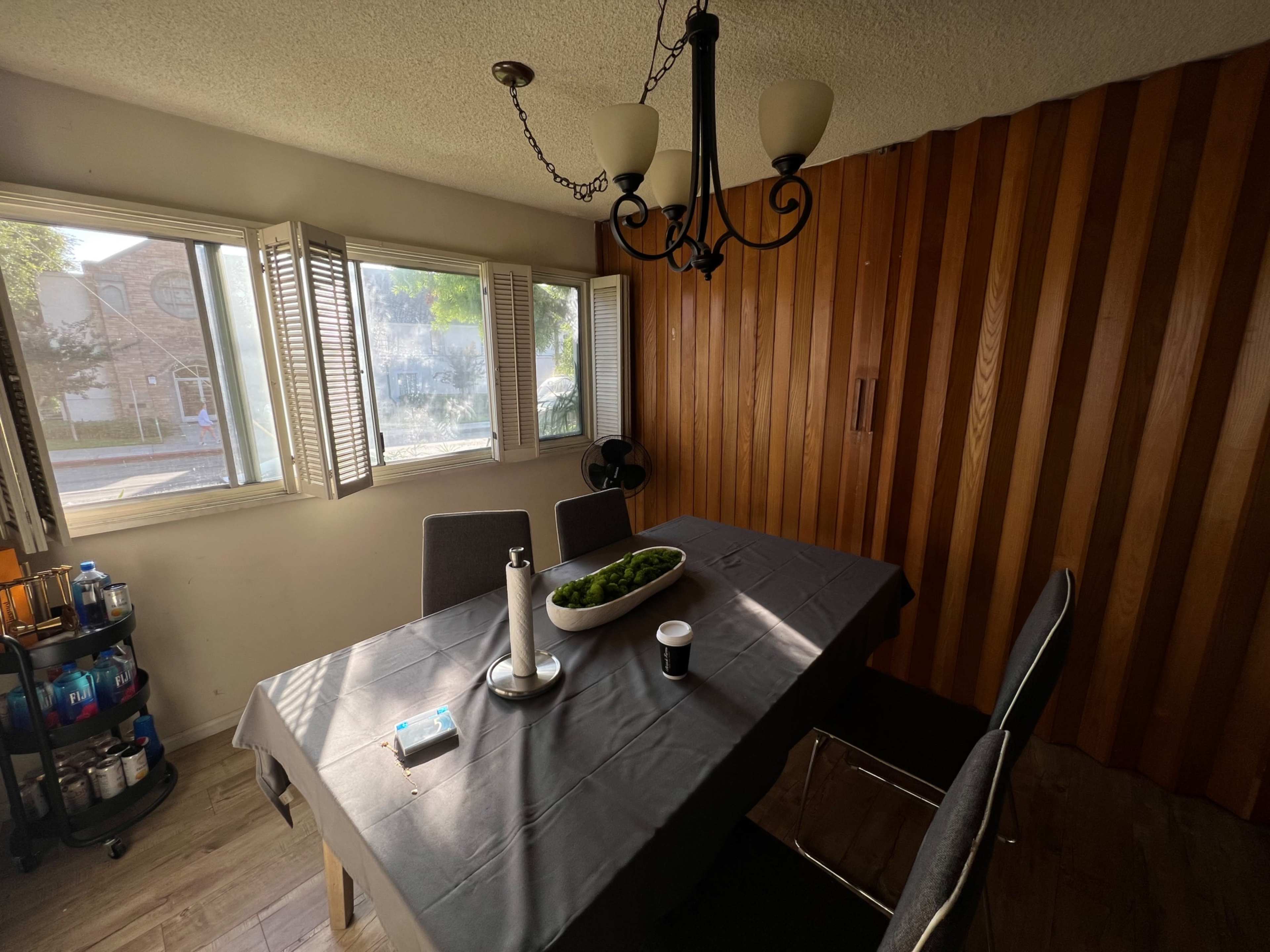 A dining room features a rectangular table set with a dark tablecloth, surrounded by gray chairs, and a chandelier hangs above the table near a window with shutters.