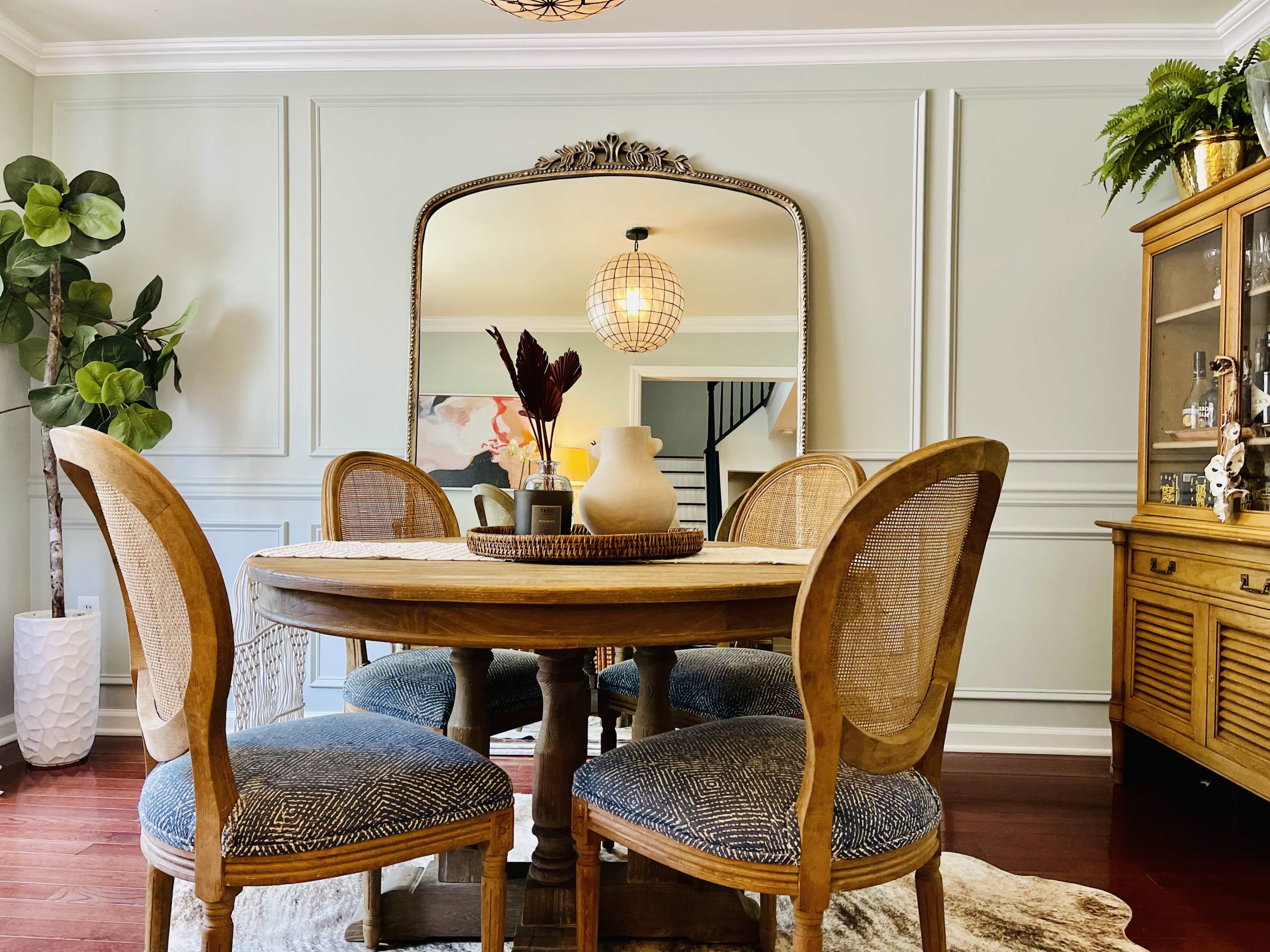 A round wooden dining table with four chairs is positioned in a well-lit room adorned with a large mirror and decorative plants.