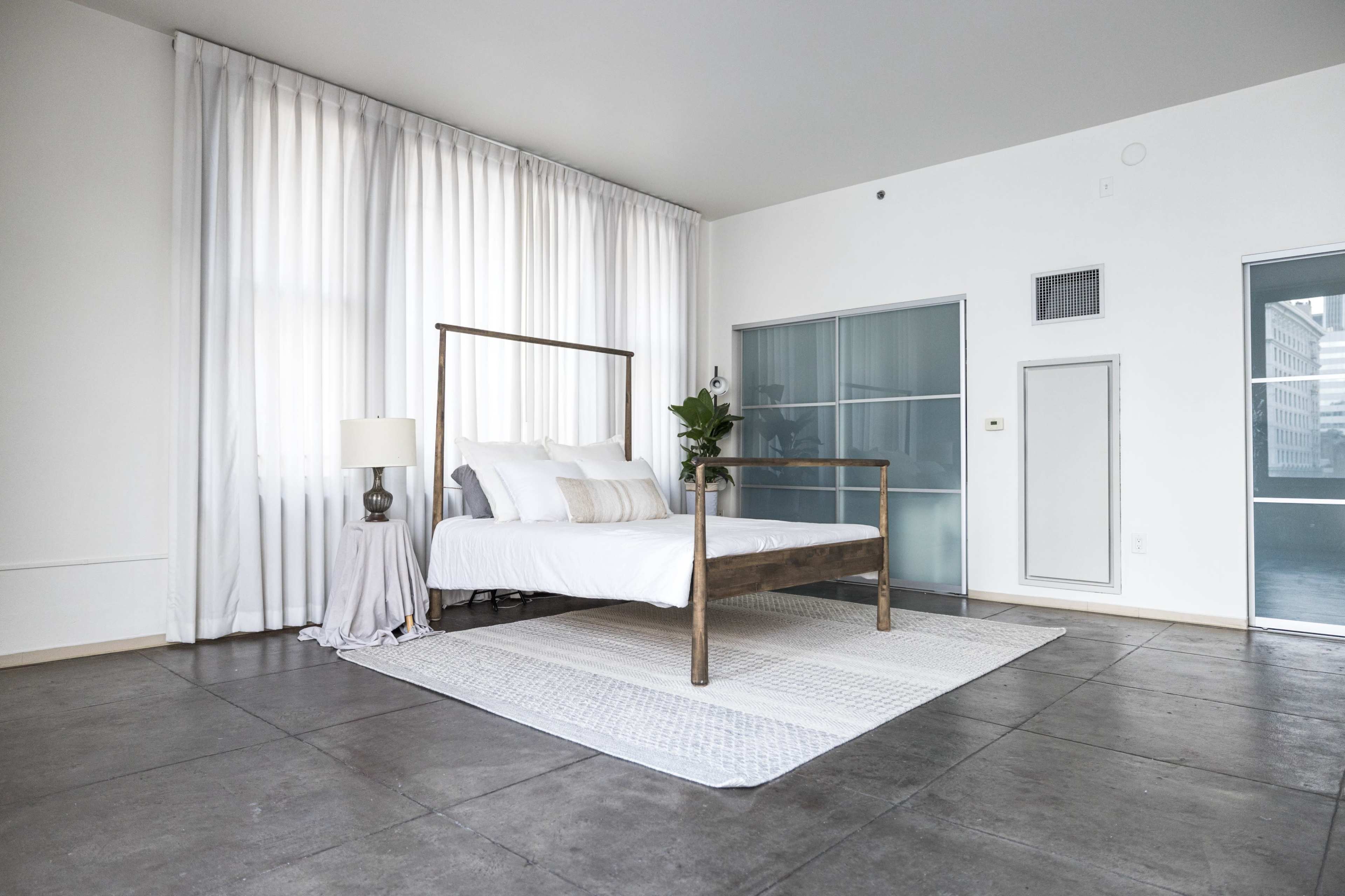 A spacious bedroom features a bed with a metal frame, white linens, a lamp on a nightstand, large windows with sheer curtains, and a concrete floor.