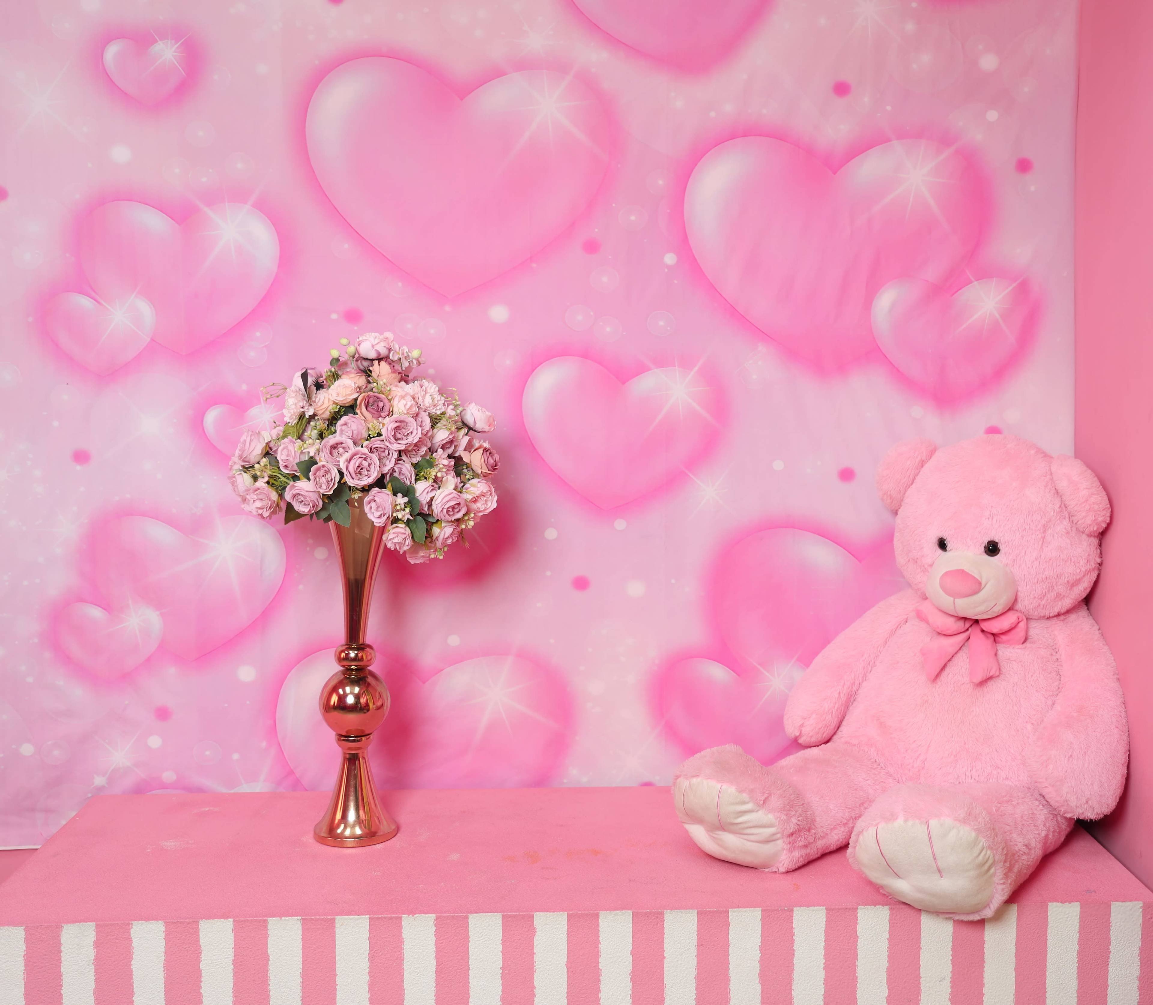 The image shows a pink-themed backdrop with a large stuffed bear seated beside a vase of pink roses on a pink platform.