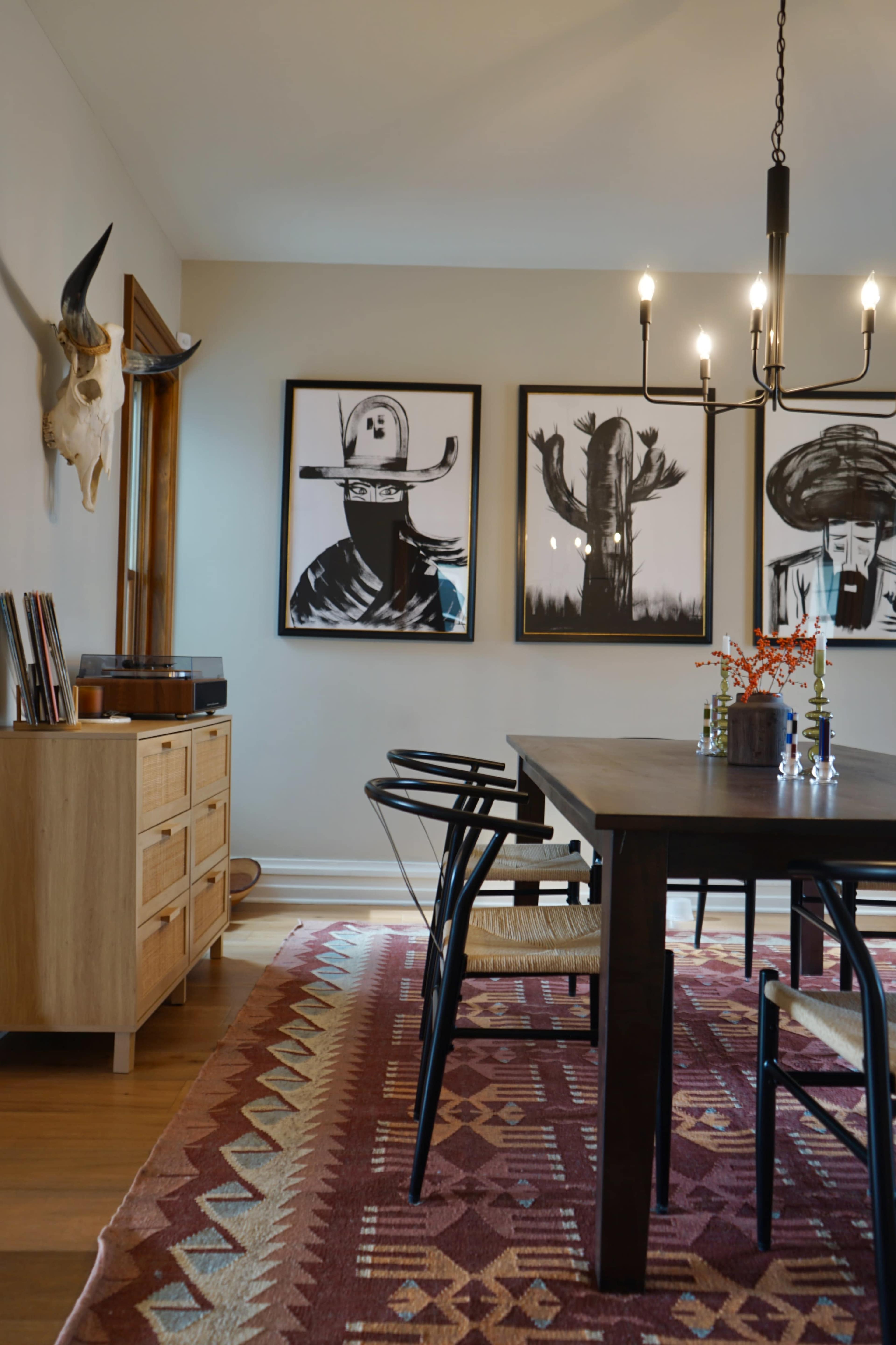 A dining room features a large table surrounded by chairs, with a patterned rug, wooden sideboard, and wall art depicting silhouettes of people and cacti.