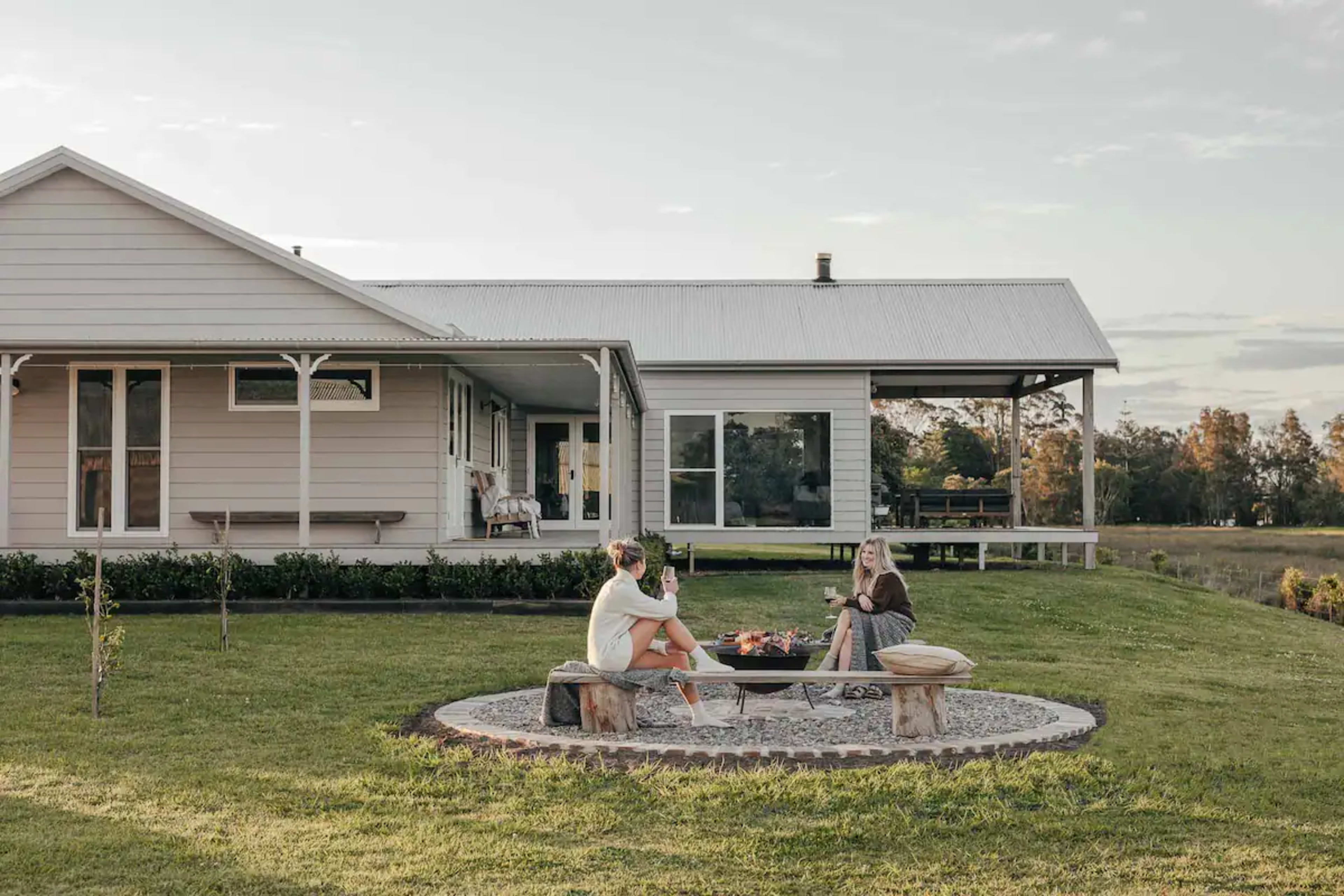 Two women sit around a stone fire pit in the yard of a light-colored house, enjoying drinks and snacks.