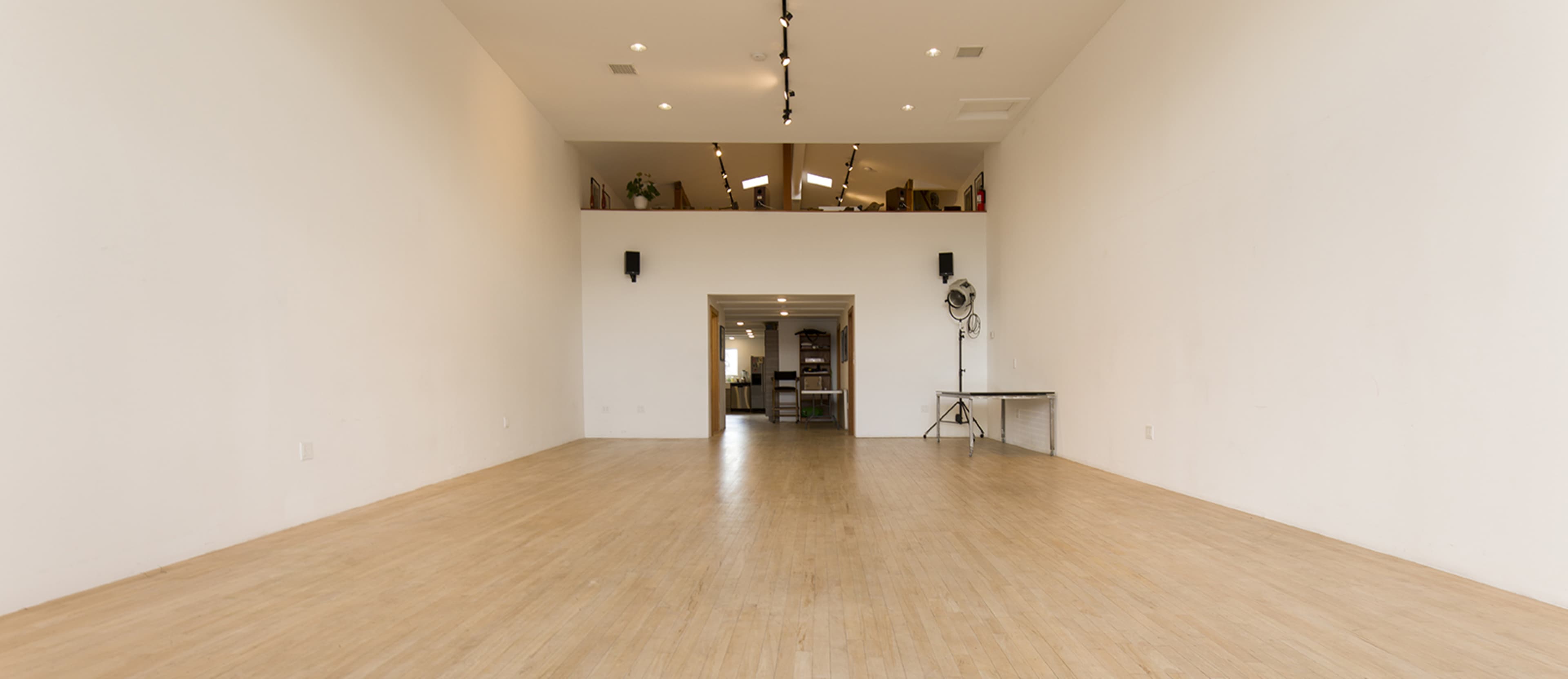 The image shows a spacious, empty indoor area with wooden flooring, white walls, and natural light coming from large windows.