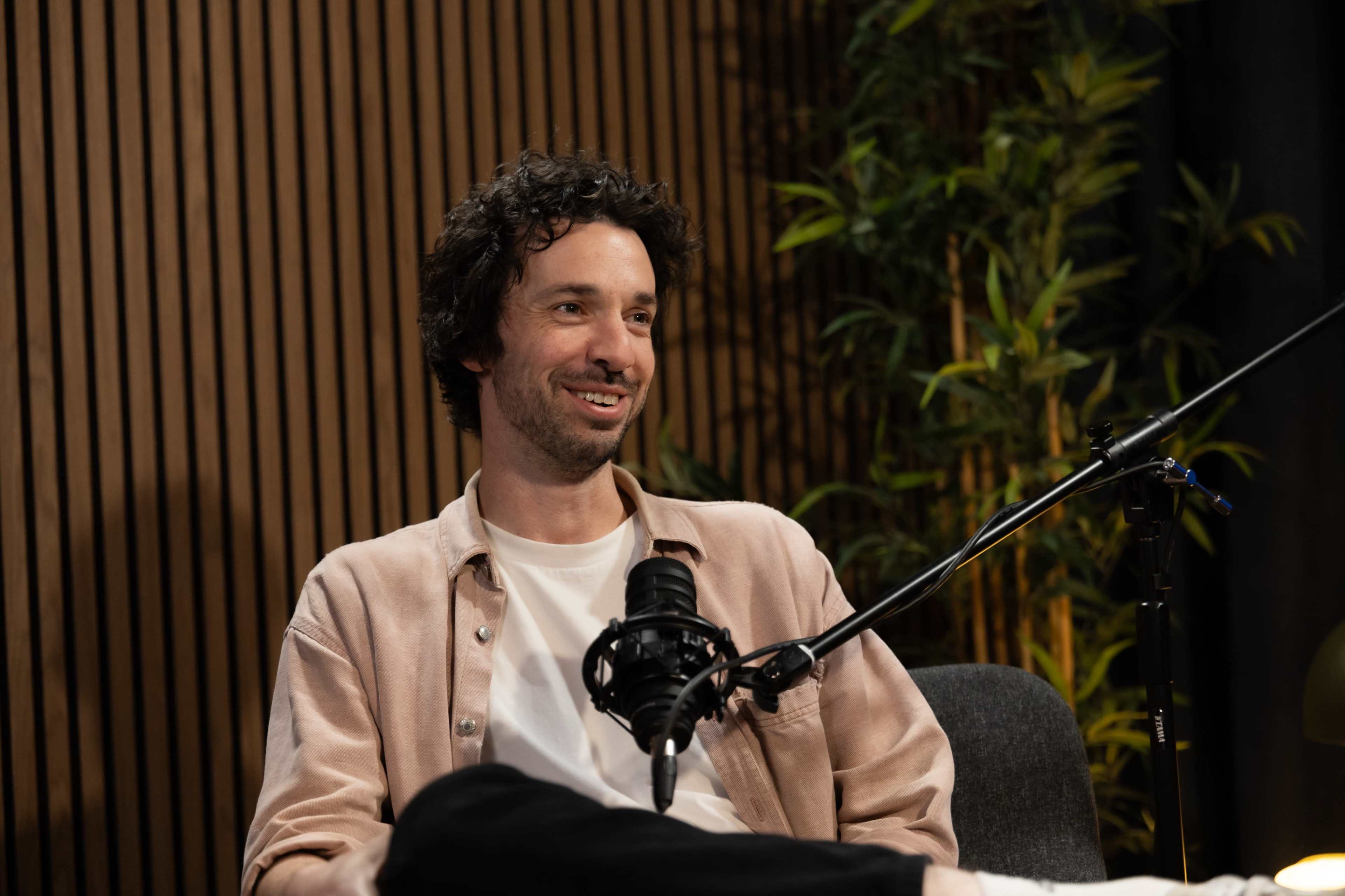 A man with curly hair is seated in a studio, smiling and wearing a light pink jacket, with a microphone positioned in front of him and greenery in the background.