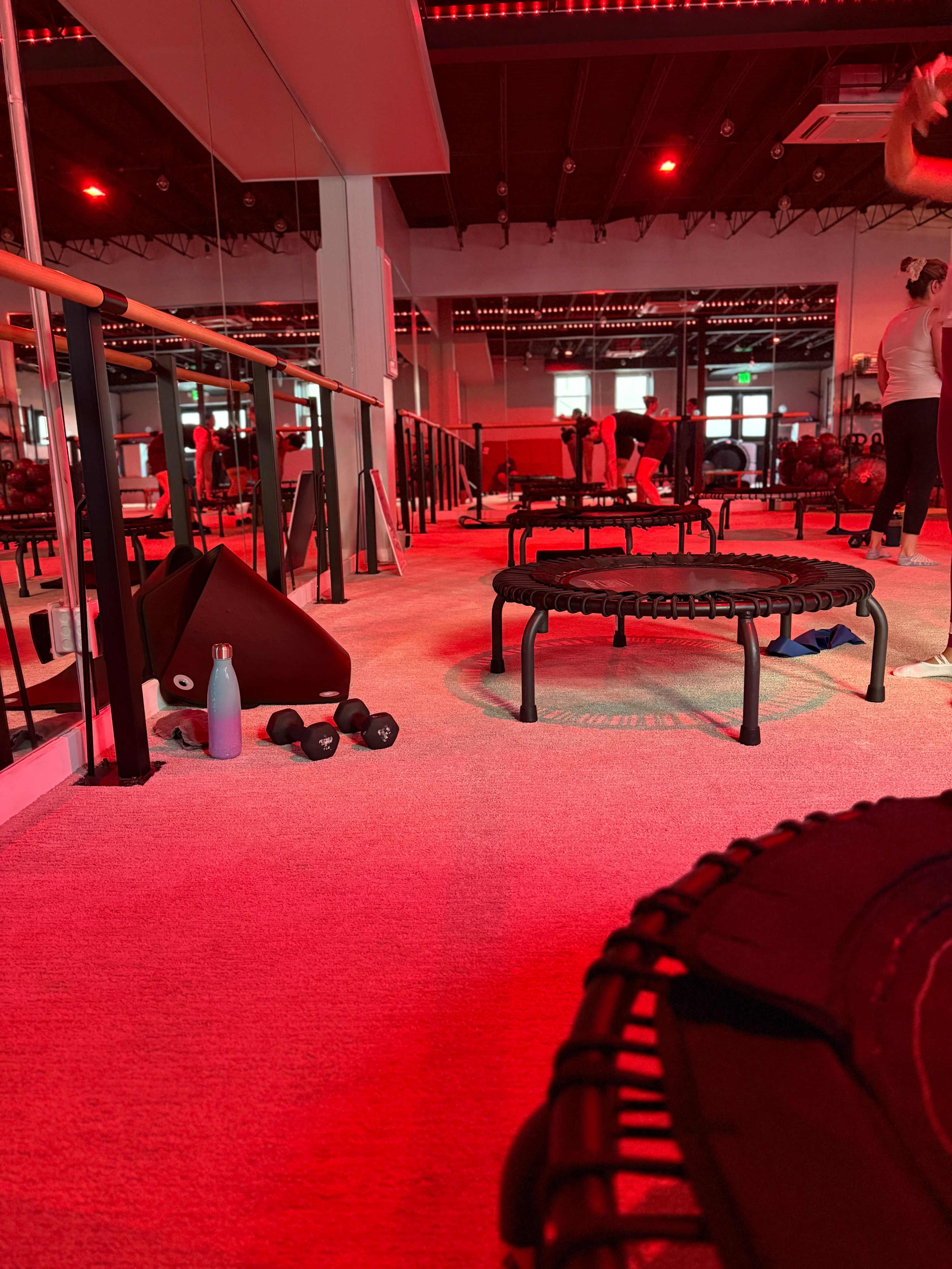 The image shows a fitness studio with red lighting, featuring exercise trampolines, dumbbells on the floor, and mirrored walls reflecting participants engaged in a workout.