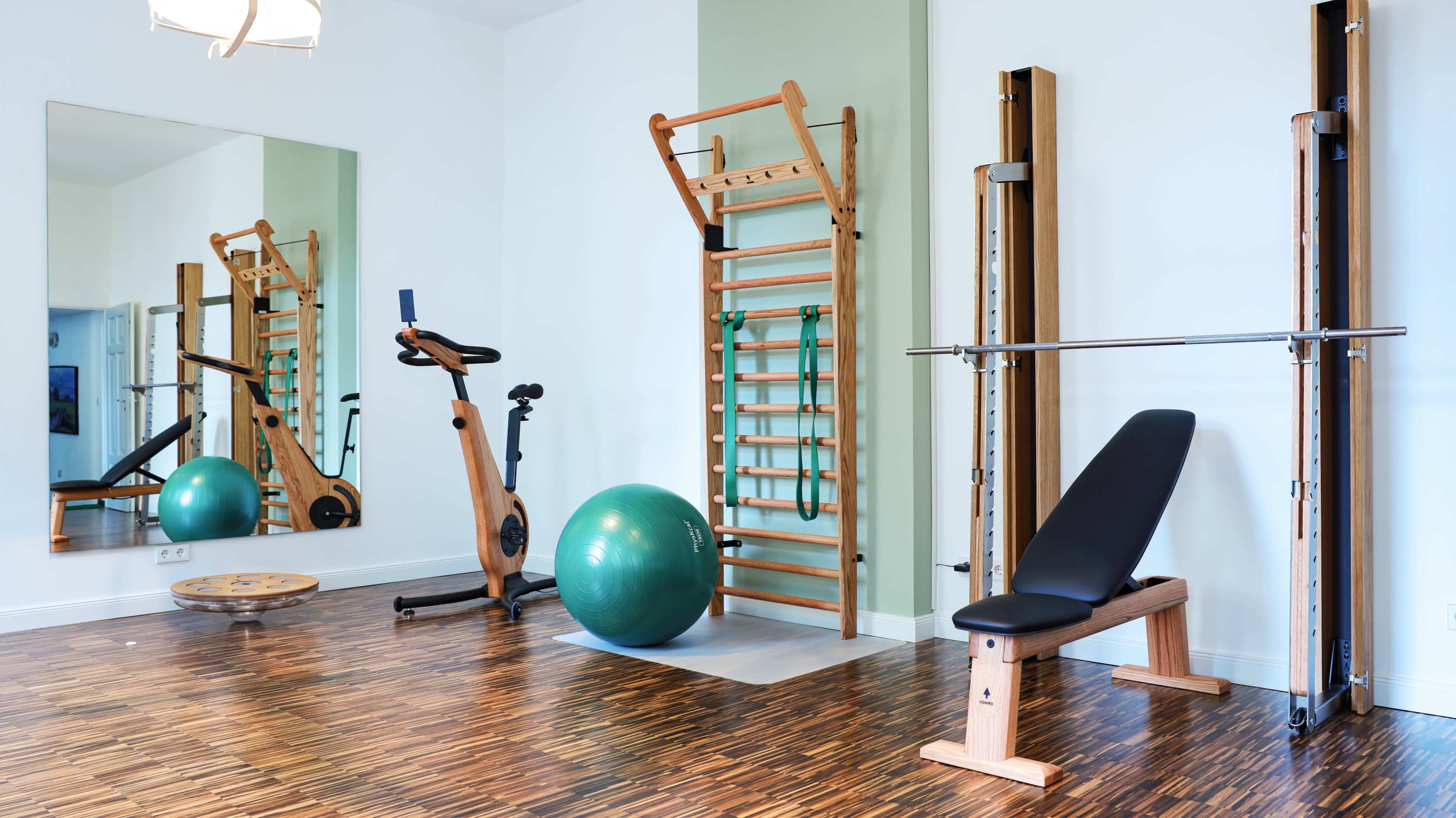 The image shows a home gym with a mirrored wall, a wooden exercise bike, a balance board, a green stability ball, a weight bench, and a vertical barbell rack against a light-colored wall.