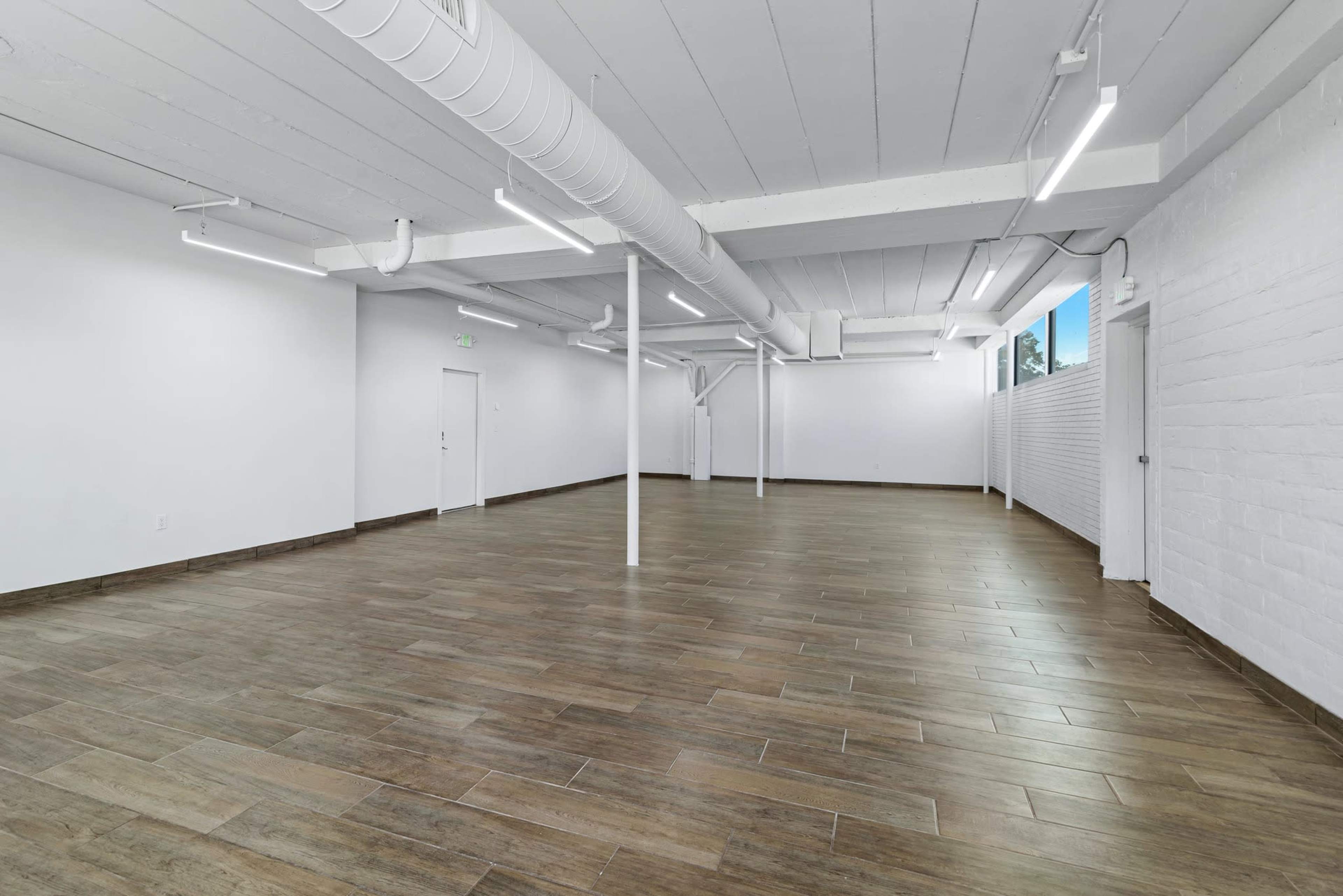 The image shows a vacant, well-lit interior space with wooden flooring and white walls.