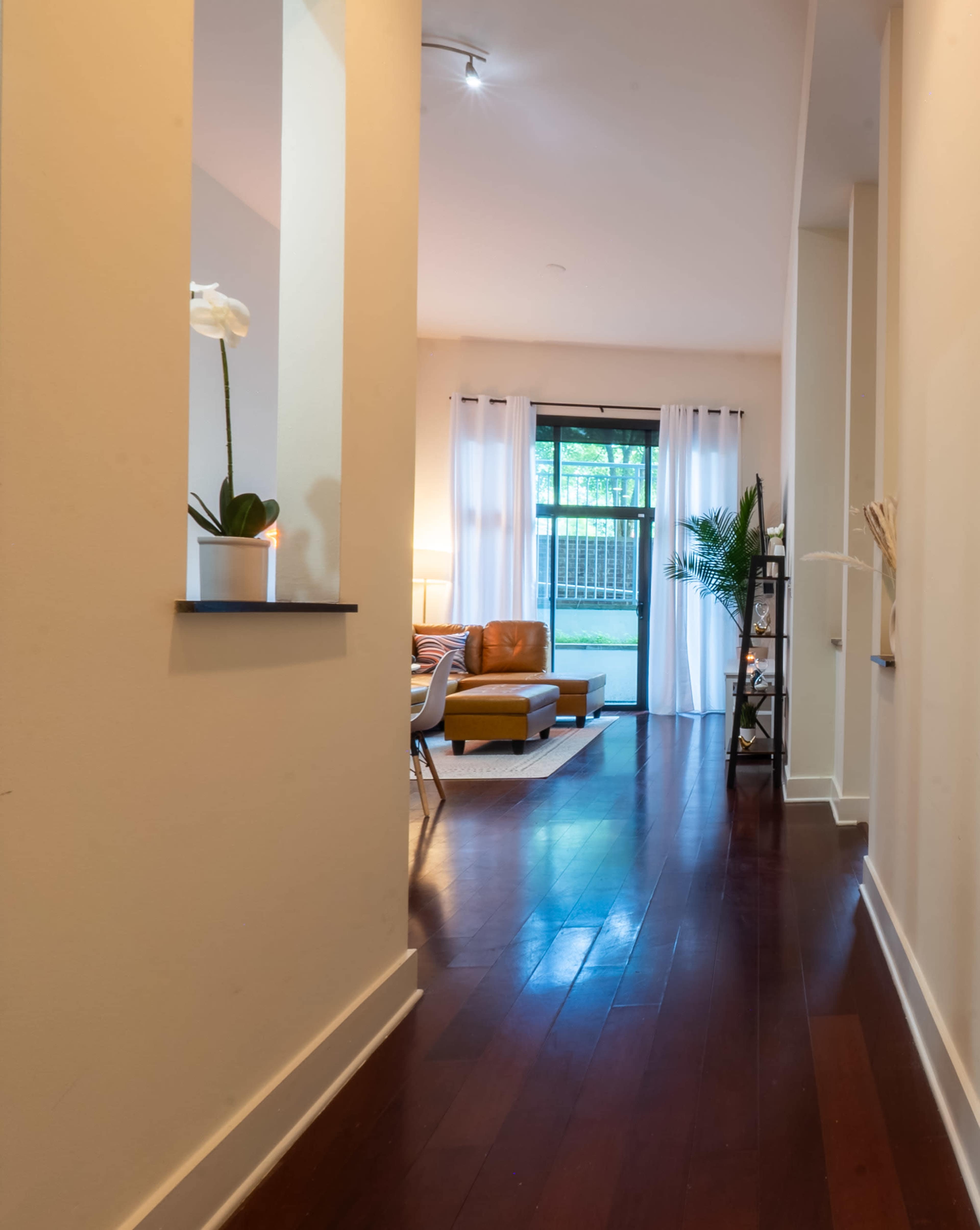 A hallway with hardwood floors leads toward a cozy living room with large windows and light-colored curtains.